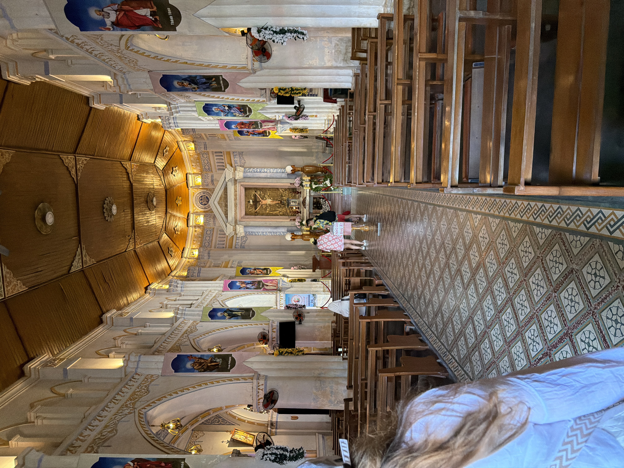 The image depicts the interior of a church, likely located in Hội Tín, Vietnam, judging by the style of the architecture and religious iconography. The perspective is from the back of the church, looking towards the altar. The church features high, arched ceilings with exposed wooden beams, ornate white columns supporting the structure, and a mosaic-tiled floor with a geometric pattern. Along the sides of the nave, rows of wooden pews are arranged for congregants.
Hanging banners with religious images and text in Vietnamese are displayed along the columns. The altar area, at the far end, is adorned with a large crucifix, religious statues, and floral decorations. Several people are visible within the church: a woman in a light-colored dress walks near the altar, a person wearing a white head covering is seated in a pew, and another individual in a pink dress is walking down the aisle. A person with long, brown hair in the foreground appears to be looking towards the altar. The overall atmosphere is one of solemnity and reverence, characteristic of a place of worship.