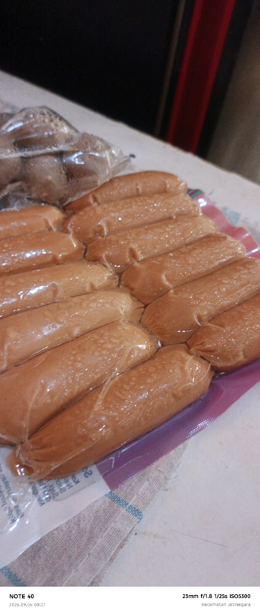 The image shows a close-up view of several rows of packaged sausages. The sausages are a uniform reddish-brown color and appear to be vacuum-sealed in plastic packaging. The packaging is somewhat translucent, allowing the texture of the sausages to be visible, with some condensation or liquid present inside. The sausages are arranged in parallel rows, with the ends of each sausage crimped. The background is out of focus, but appears to be a surface with some texture, possibly a table or counter. The bottom of the image contains text overlay, including "NOTE 40," a date and time "2026.09.06 08:21," and camera settings "23mm f/1.8 1/25s ISO5300," along with the location "Kecamatan Jatinegara," which is a subdistrict in East Jakarta, Indonesia.