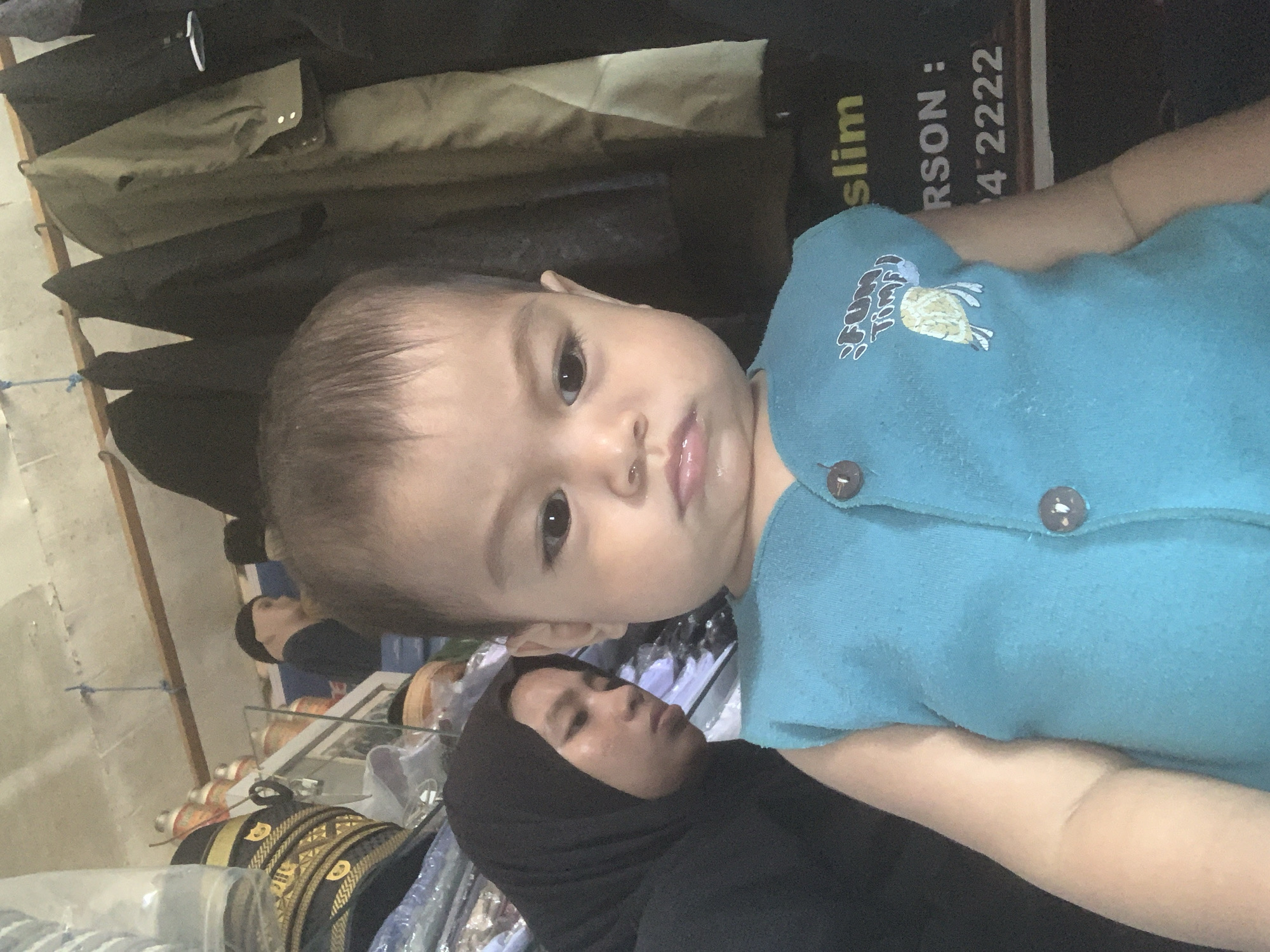 The image, rotated 90 degrees clockwise, presents a close-up of a baby/toddler held by an adult woman, likely in an indoor setting such as a shop or market stall in Sungai Meriam, Indonesia.
The baby, appearing to be a toddler, is prominently featured in the foreground, looking directly at the camera. They have light brown hair, dark eyes, and a neutral, slightly inquisitive expression. The baby is wearing a light blue collared shirt with two brown buttons. On the shirt, a cartoon-like graphic depicts a bird, with partially visible yellow text that appears to read "S FUN TOWN."
Behind and below the baby, an adult woman is visible, wearing a black hijab and black clothing. Her face is partially obscured by the baby, and her gaze appears to be directed downwards or away from the camera.
The background shows elements consistent with a retail environment. To the left, a clothing rack holds various dark-colored garments. On the right side, a black sign or banner with yellow lettering is visible. Legible text fragments include "slim," "RSON :," and "4 2222," which could be part of a business name or contact information. Below this sign, there are hints of more clothing or textile items. Further back, a counter or display case holds assorted items, including what looks like rolls of decorative trim or fabric, some with gold and black patterns. The lighting suggests an indoor daytime scene.
