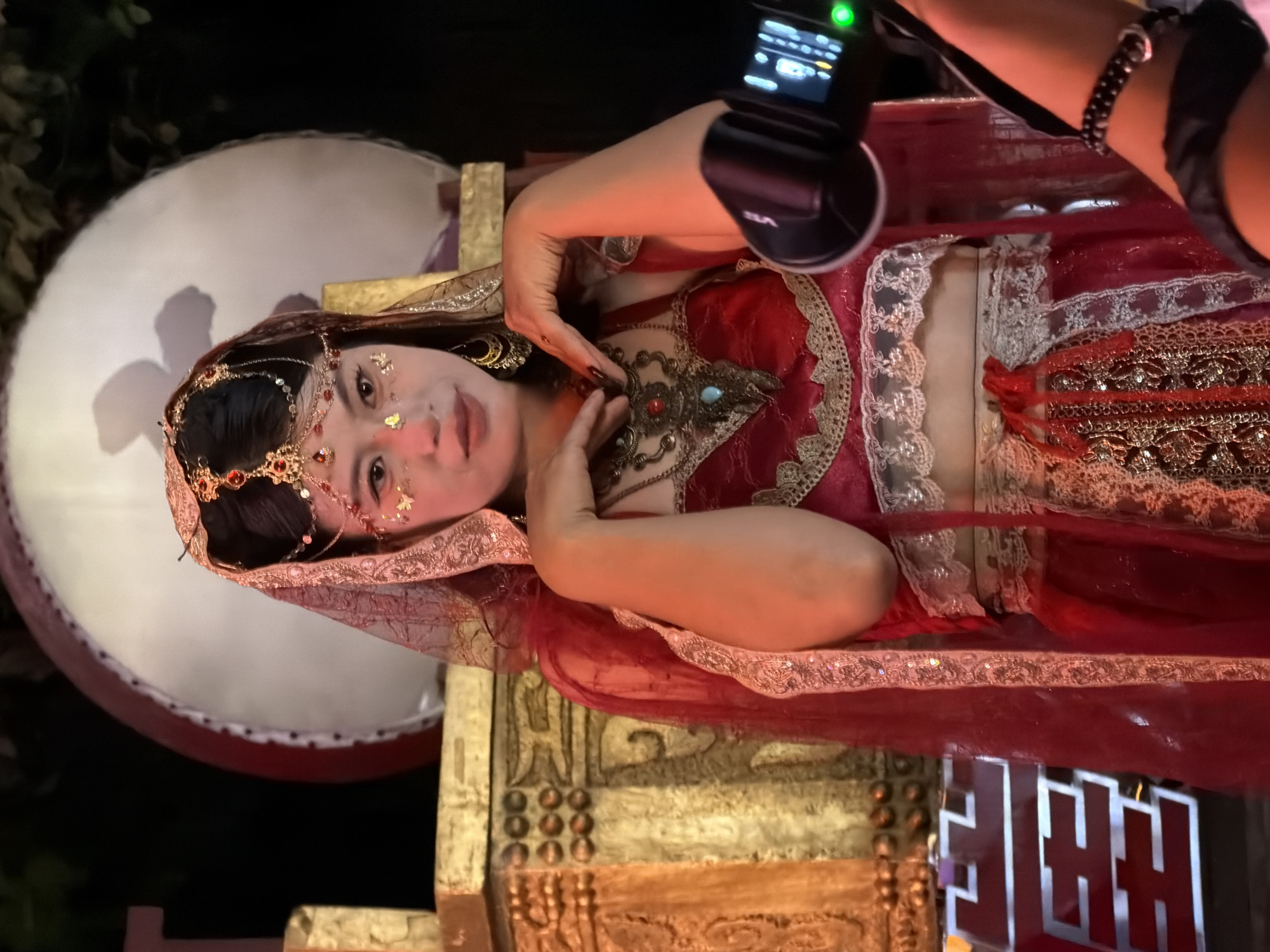 A woman in traditional Indian attire is posing for a photo in front of a large drum and ornate golden structures. She wears a red veil, a beaded headdress, and elaborate jewelry. The background appears to be a night scene, possibly an outdoor event or performance, with a large white circular object that resembles a drum with a red rim. The woman is positioned in the center of the frame, with her hands near her face, and her expression is serene. The golden structures to her left are intricately carved and appear to be part of a set or stage. In the foreground, a camera or recording device is visible, with a hand holding it. The overall scene suggests a cultural performance or photoshoot. The location is identified as Yuzhong District, China.