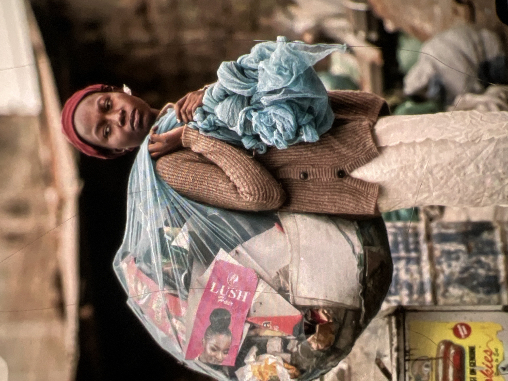 A young person, likely a woman, is shown from the chest up, carrying a large, clear plastic bag of refuse over their right shoulder. She wears a red head covering and a brown knitted cardigan over a white garment. A blue cloth is draped over her left shoulder and hand. The bag contains various items, including what appears to be a pink box with text and an image. The background suggests an urban environment, possibly a street or alleyway in Lagos, Nigeria, with debris and structures visible. The lighting is somewhat diffused, with hints of bright sunlight. The image has a textured, possibly aged or filtered appearance.