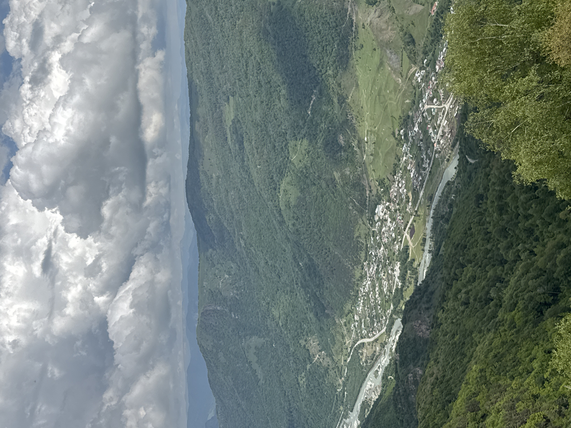 The image captures a panoramic view of a valley with a village nestled within it, bordered by a river and flanked by densely forested mountains. The sky is filled with dramatic, heavy clouds, suggesting an overcast day. In the foreground, lush green trees, likely birch trees, dominate the bottom right of the frame, with their light green foliage reaching upwards. The middle ground reveals the village of Nizhnyaya Teberda, Russia, characterized by numerous small houses with red and gray roofs, spread out along a road that follows the winding path of a river. The river itself is wide and appears to have a rocky bed, with its water reflecting the muted light of the sky. The background consists of rolling, verdant mountains covered in thick forests, their slopes crisscrossed with visible roads or paths. In the distance, further mountain ranges are visible, their forms softened by atmospheric haze. The overall scene conveys a sense of remote, natural beauty and a settled, rural community within a dramatic mountainous landscape.