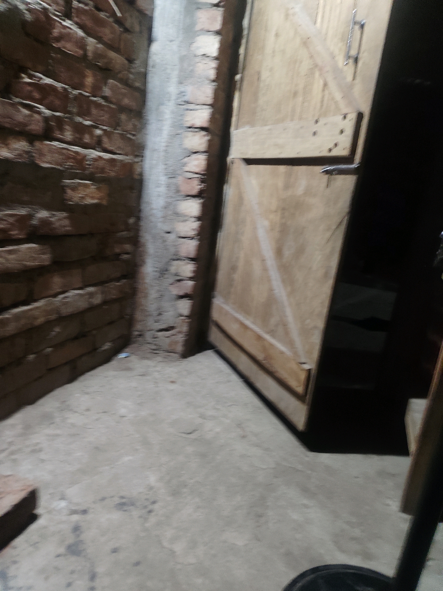 The image depicts an interior corner of what appears to be a dimly lit room or corridor. To the left, a rough brick wall is visible, constructed from reddish-brown bricks. Adjacent to this wall is a vertical concrete pillar, also showing rough texture. To the right, a wooden door is positioned slightly ajar. The door is made of light-colored planks and has a visible handle and latch mechanism. The floor is concrete, showing some unevenness and scattered dark marks. The lighting is dim, casting shadows and obscuring details in the background to the right of the door. There are no people or animals present in the frame. The overall scene suggests an unfinished or utilitarian space. The location in Amdai, India is not discernible from the visual information provided.