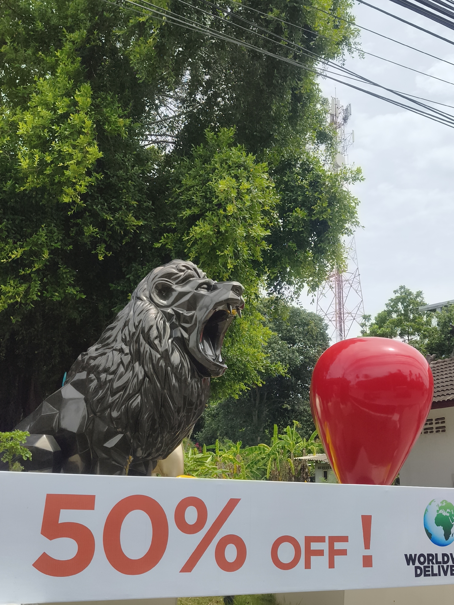 A horizontal, medium shot captures an outdoor scene in the city of Ko Samui District, Thailand, featuring a large, dark, angular sculpture of a roaring lion in the left foreground. To the right of the lion sculpture, a bright red, heart-shaped object is prominently displayed. In the lower portion of the image, a white banner with large, orange lettering reads "50% OFF!". To the right of this text, a small graphic of the Earth with a green continents and blue oceans is visible, accompanied by text that begins to read "WORLDWI DELIVE". Behind the lion and the heart, lush green trees dominate the background, interspersed with a tall, red and white lattice telecommunications tower and overcast sky. A light-colored building with a tiled roof is partially visible on the far right. The overall scene appears to be an advertisement or a storefront display.