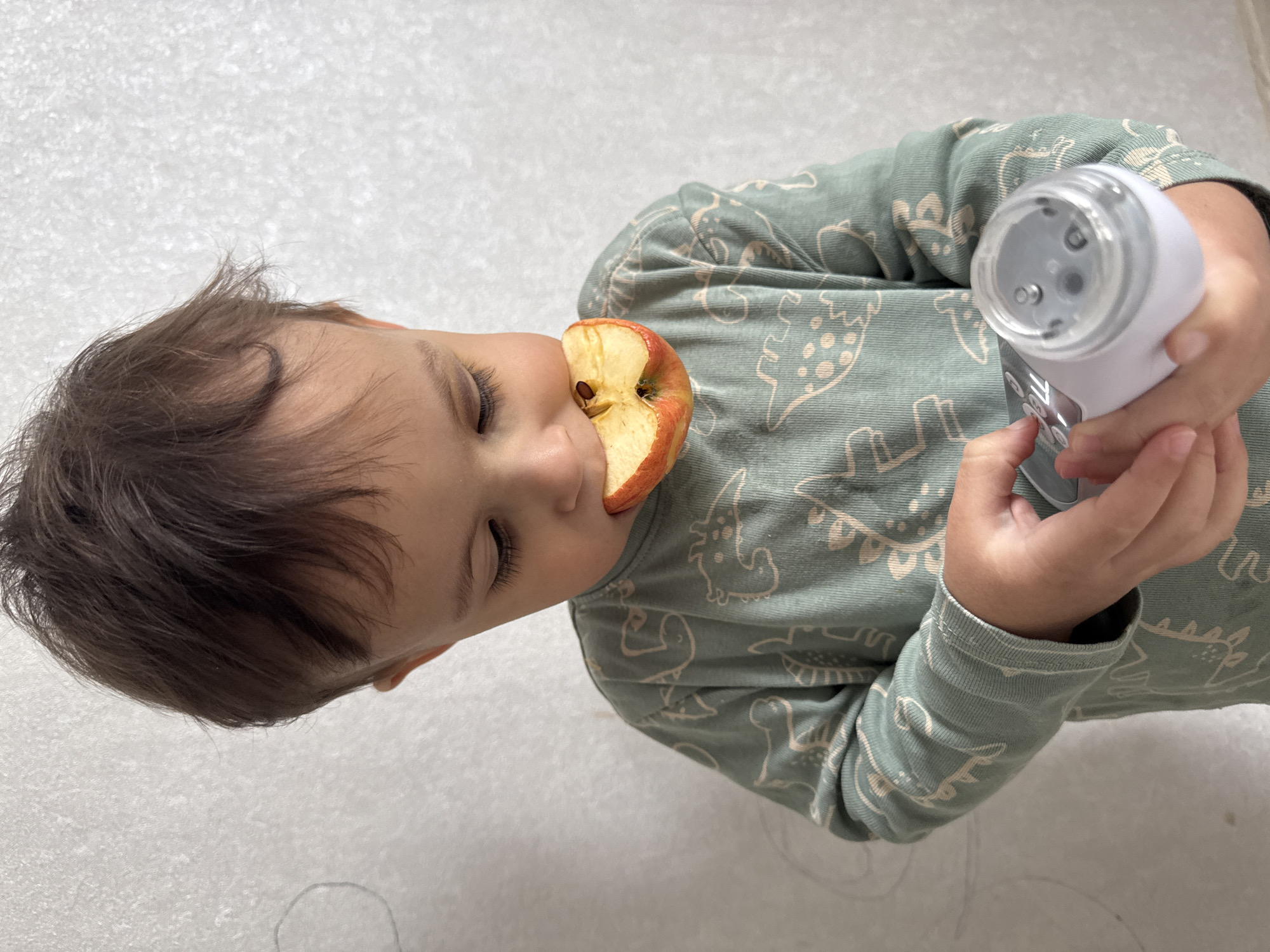 A child is holding a white and grey electric nail file in their right hand and an apple slice in their mouth. The child has brown hair, closed eyes, and is wearing a green long-sleeved shirt with a dinosaur pattern. The background is a plain, light grey or white surface. The photograph may have been taken in Chaikovsky, Russia, but the scene provides no location-specific details.