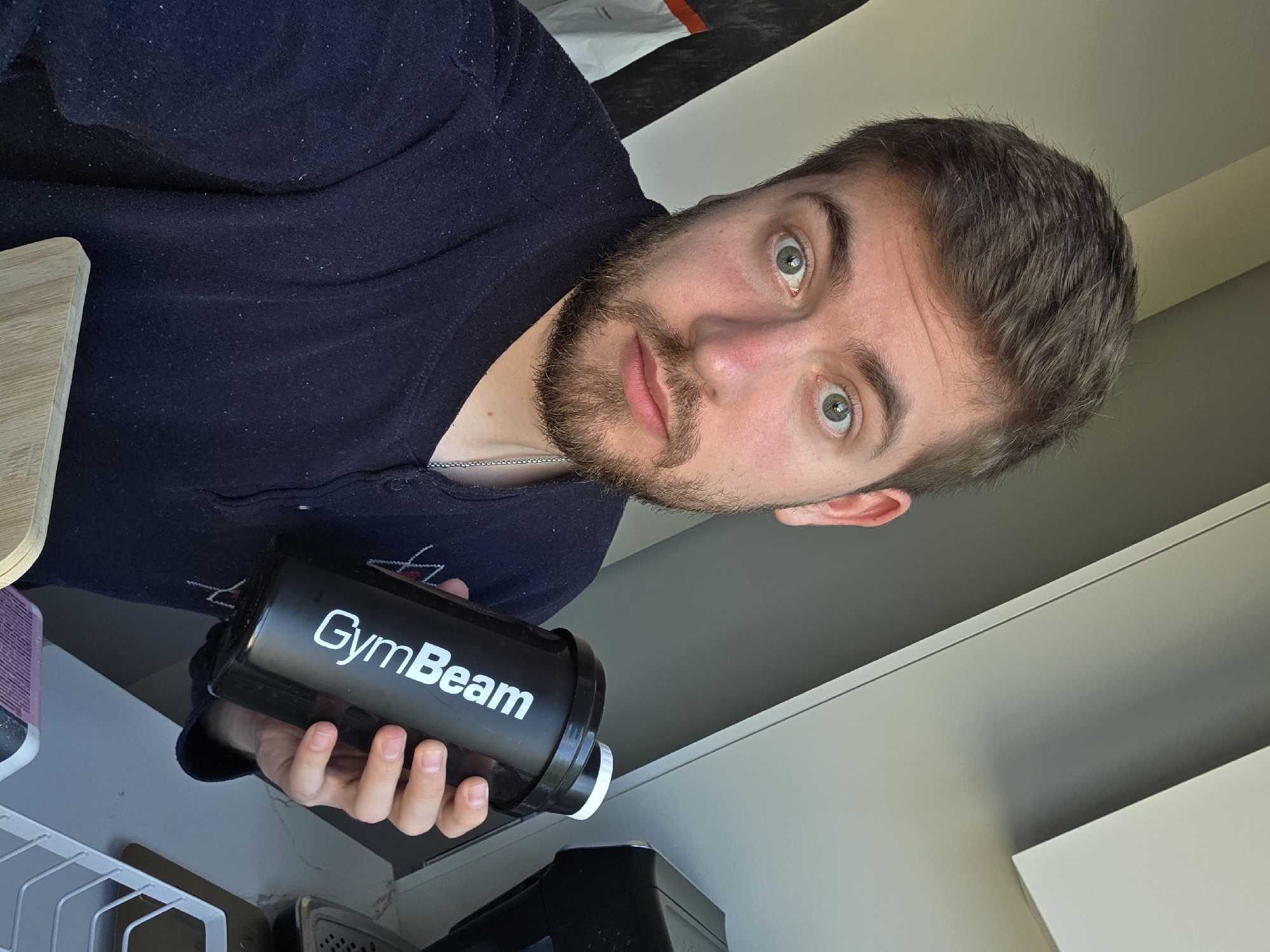 A man is holding a black GymBeam shaker bottle. The man has short brown hair, green eyes, and a beard. He is wearing a dark blue shirt. The background appears to be a kitchen setting, with a light-colored countertop and cabinets visible. It is not possible to determine the location in the city of Yagodovo, Bulgaria, from the image.