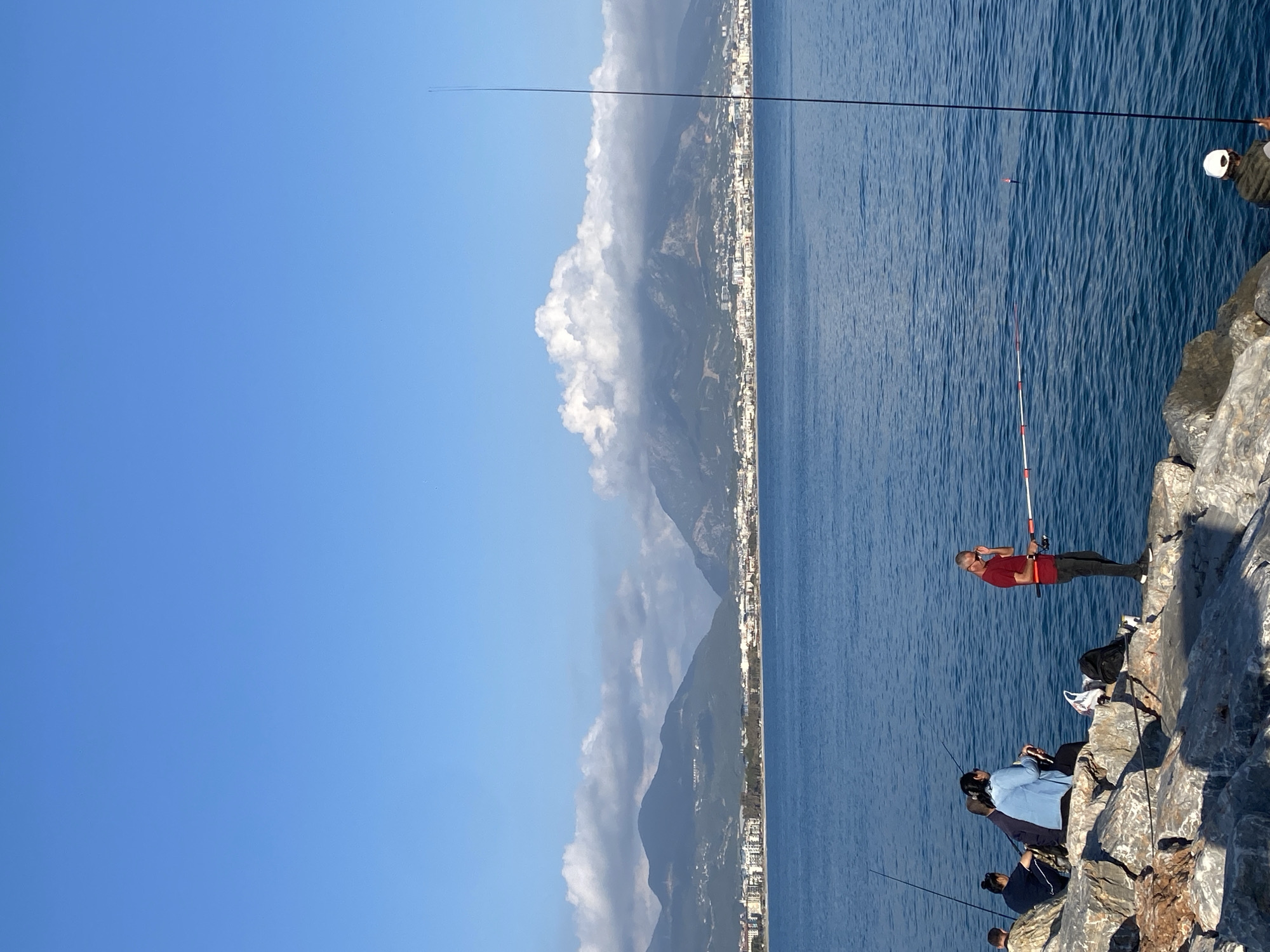 The image depicts a vibrant coastal scene in Alanya, Turkey, on a clear and sunny day. The foreground showcases a rugged rocky shoreline, serving as a platform for several individuals engaged in fishing.
A prominent figure, a man in a red shirt and dark pants, stands on a large grey rock, intently holding a fishing rod extended over the deep blue sea. Below him, a small group of people are seated or standing on the lower rocks, also participating in the activity with their fishing rods cast into the water. One person is visible in a light blue top, while another wears dark clothing. A white-hatted individual is partially seen in the upper right, also fishing. A red and white fishing bobber floats in the calm water, indicating active fishing. A black backpack and some lighter-colored items are placed on the rocks near the man in the red shirt.
The expansive blue sea occupies a significant portion of the left side of the frame, exhibiting subtle ripples. In the distance, a long stretch of coastline is visible, featuring undulating mountains partially obscured by scattered white and grey clouds. Along the base of these mountains, a densely built-up urban area with numerous structures lines the shore. The sky above is a bright and cloudless blue. The overall ambiance is one of leisure and tranquility, capturing a typical day of seaside fishing. No discernible text is visible in the image.