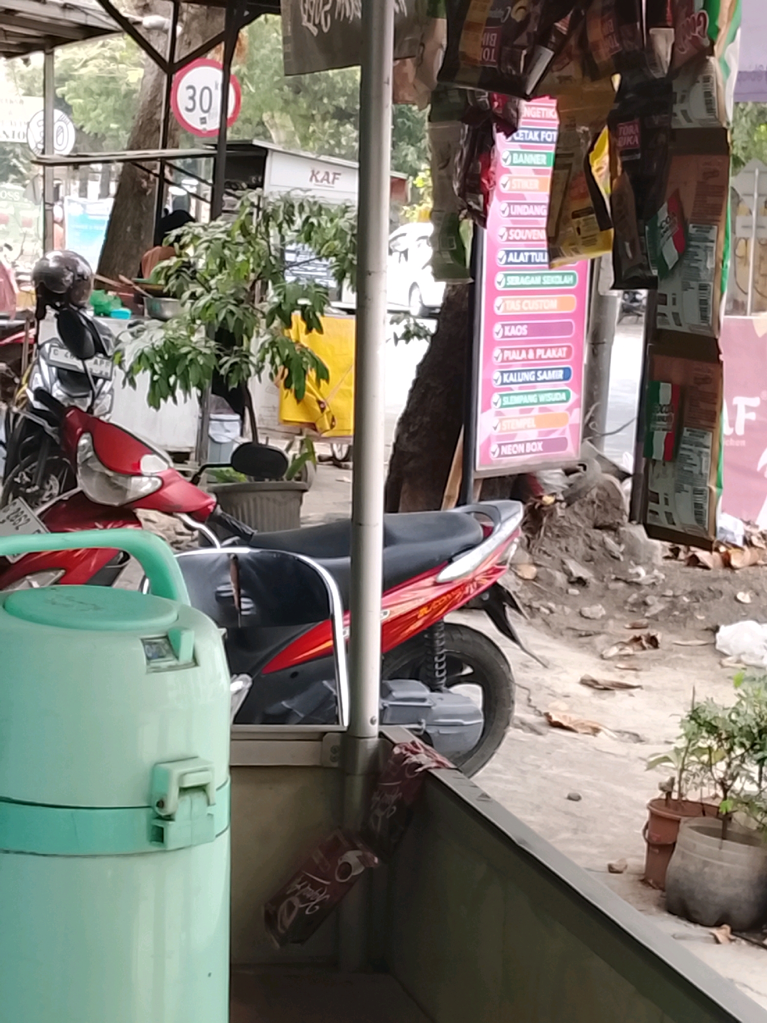 The image depicts an outdoor scene in Slawi, Indonesia, likely a roadside stall or shop. In the foreground, a light green cylindrical object with a teal band and a lid is visible on the left. To the right of this, a red and black motorcycle is parked, with its front wheel obscured. Behind the motorcycle, a potted plant with green leaves sits on the ground.
Further back, a sign with various services listed in colorful boxes, such as "CETAK FOTO," "BANNER," "KAOS," and "NEON BOX," is mounted on a wooden pole. Several packets of snacks or other goods are hanging from a structure above this sign. A "30" speed limit sign in a red circle is also visible on a pole. In the background, trees and a white building can be seen. The ground appears to be paved with concrete, with some scattered leaves and debris. The overall impression is one of a functional, possibly small business operating in a semi-urban environment.