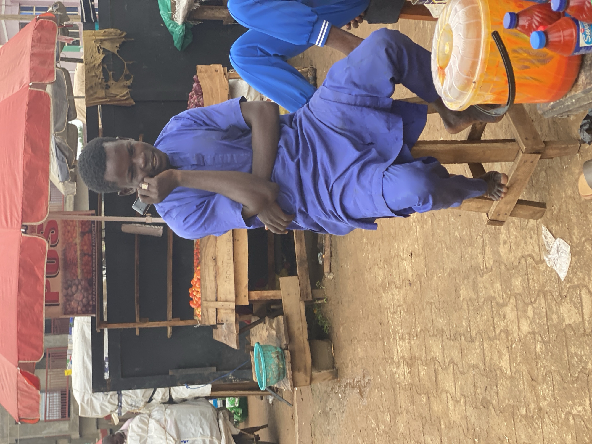 The image captures a man in Kafanchan B, Nigeria, engaged in a phone conversation while seated on a wooden structure, possibly a makeshift bench or stall. He is dressed in a blue, flowing garment. The scene is an outdoor market environment with stalls selling various goods. To the man's right is a wooden platform holding a covered orange bucket and several bottles of what appears to be a bottled beverage. In the background are stalls and various produce, including tomatoes. Underneath, in front, and behind the stalls are bags, tarps, various buckets, and waste debris that is found in open-air market settings. Overhead are red awnings. The ground is patterned with what appears to be concrete. A sign for the Kafanchan Modern Market is partially visible. Another person is seated above the man in a similar-style blue garment with white-striped pants.