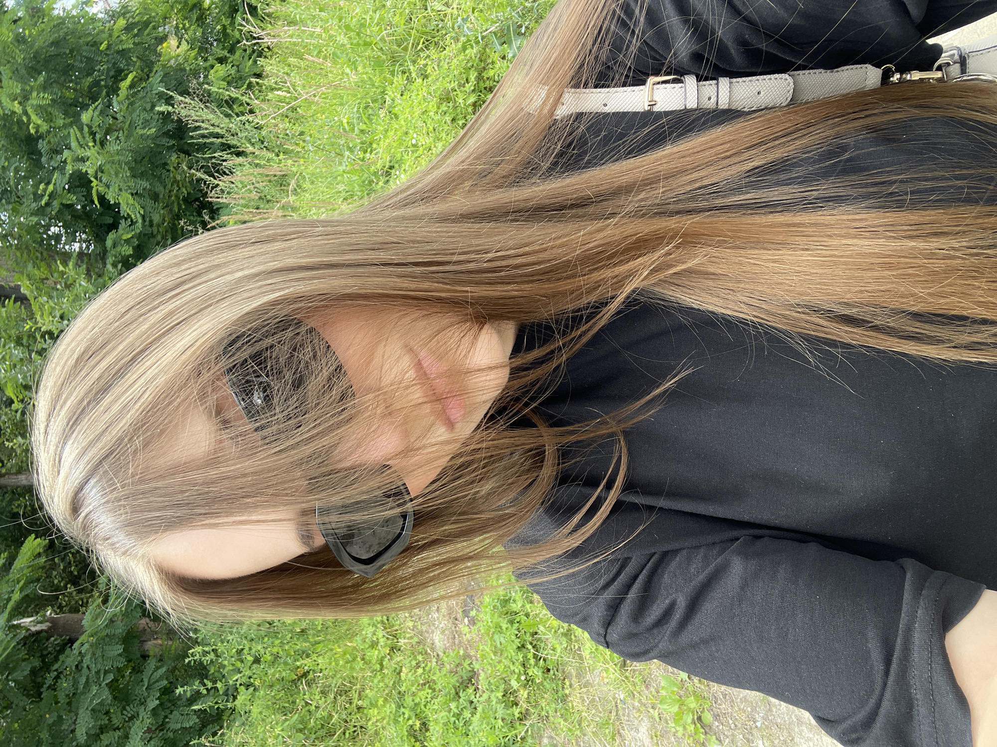A young woman with long, light brown hair and sunglasses is taking a selfie outdoors. She is wearing a black long-sleeved shirt. A light-colored purse strap is visible over her right shoulder. The background consists of green trees and foliage, suggesting a park or natural setting. The image appears to be taken in Konopnytsia, Ukraine, based on the provided context.