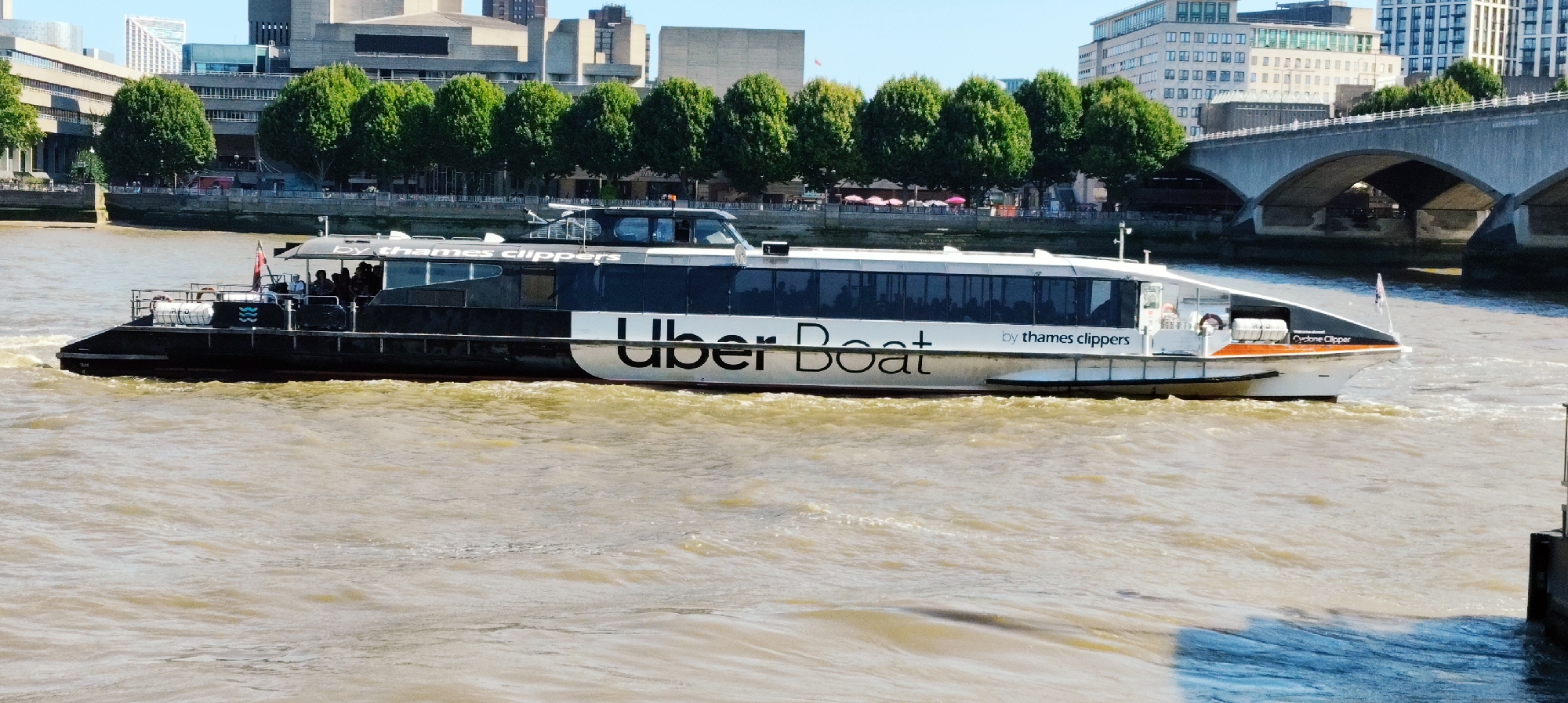 An Uber Boat by Thames Clippers ferry is moving rightward on a muddy river in London, United Kingdom. The ferry is white with black and dark blue accents. The words "Uber Boat" are prominently displayed in large white lettering on the side of the ferry, with "by thames clippers" written in smaller text above it. Several people are visible inside the ferry, and some are standing on the open deck at the rear of the vessel. The river is choppy with ripples and waves, suggesting movement. In the background, a large, arched concrete bridge spans the river. Behind the bridge, there are buildings, including some modern office buildings with a prominent glass facade. Trees with dense green foliage line the riverbank between the ferry and the buildings. The sky is clear and blue, indicating a sunny day. The overall scene depicts urban transportation on the River Thames.