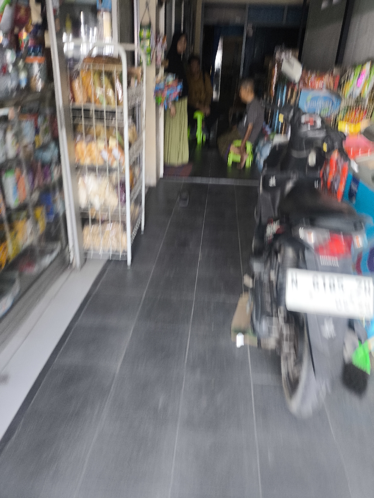 The image depicts the exterior of a small store or warung in Lumajang, Indonesia. The foreground is dominated by a dark gray tiled floor. To the left, shelves stocked with various packaged goods are visible through a glass enclosure. A white metal display rack with multiple shelves, filled with bagged snacks and food items, stands in the middle ground. A black motorcycle is parked in the right middle ground, with its license plate partially visible. In the background, through an open doorway, several people are present. One individual is wearing a dark headscarf and a light-colored garment, holding a bag. Another person, wearing a dark t-shirt, is seated on a green plastic chair. The overall impression is a busy, informal commercial setting in a city.