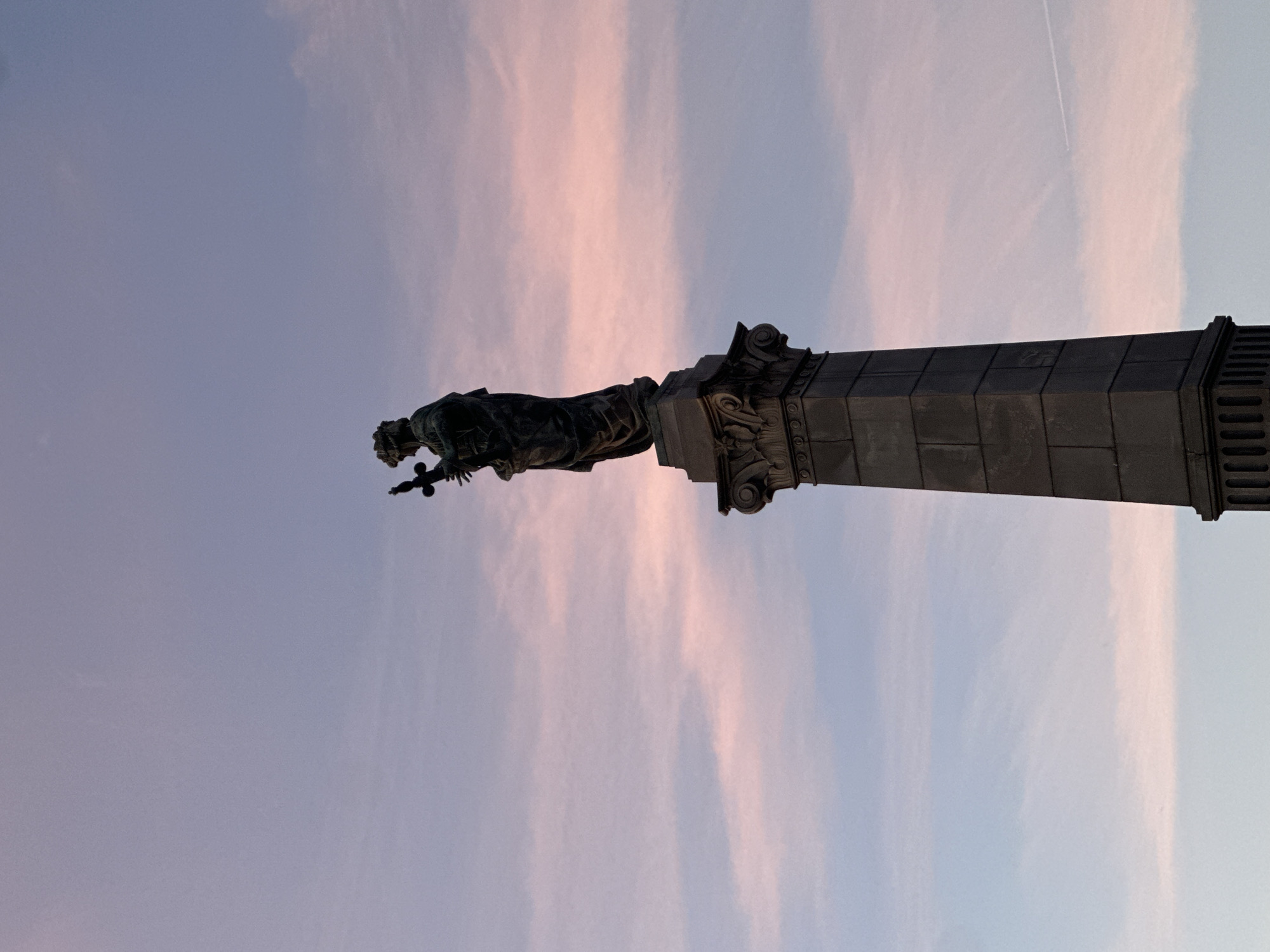 The image depicts a statue atop a column against a backdrop of a light blue and pink streaked sky. The statue, likely made of bronze or a similar dark metal, stands on an ornate pedestal with classical architectural details. The sky has a subtle gradient of color, suggesting either sunrise or sunset. This is the Statue of Liberty in Ruse, Bulgaria.