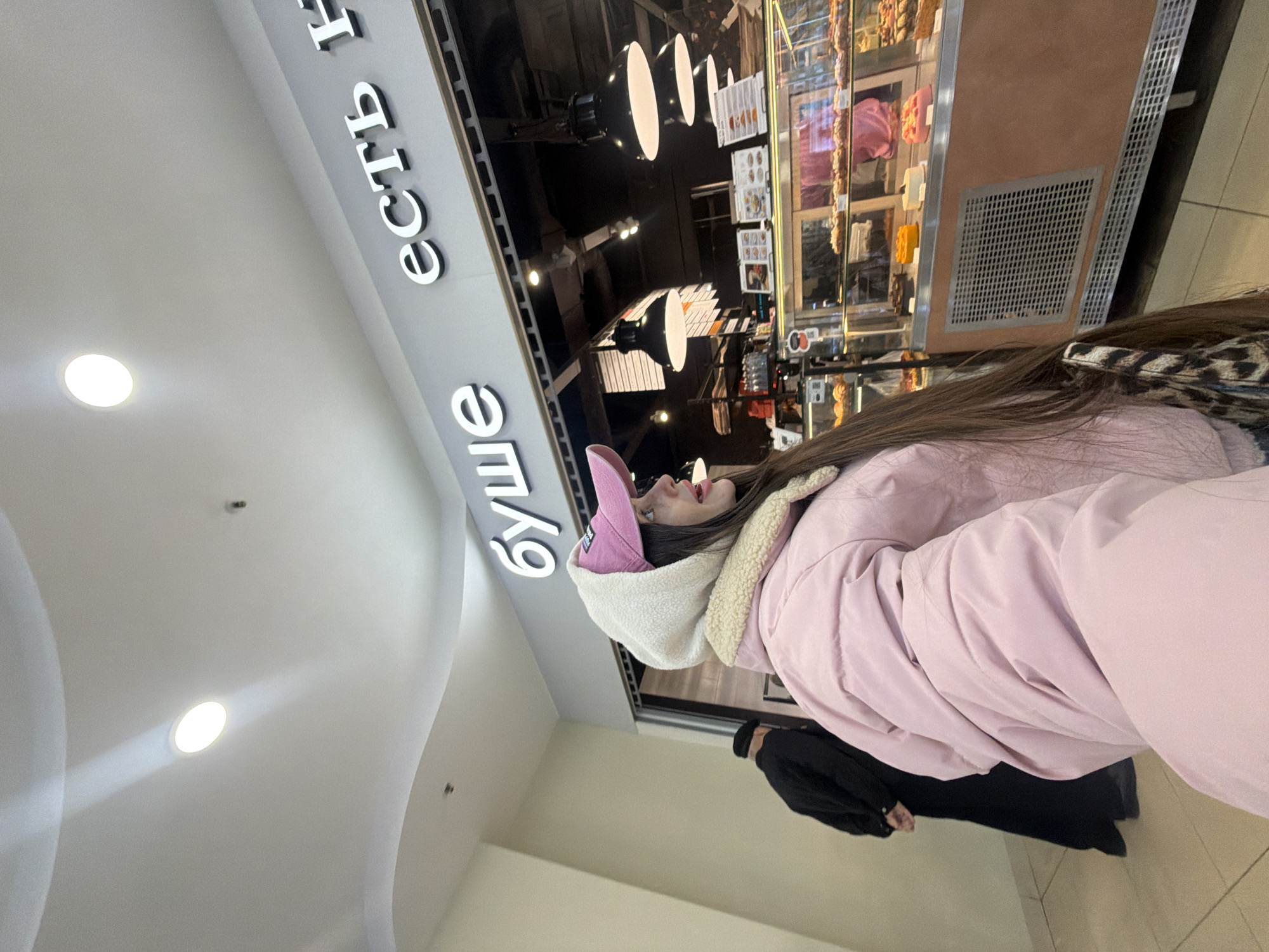 Here is a detailed description of the image:
**Scene**: The image captures a scene inside a building, likely a shopping mall or food court, in Saint Petersburg, Russia. This is indicated by the Cyrillic lettering ("буше есть") on the signage above the person.
**People**: The primary subject is a young woman wearing a pink puffer jacket with a white, sherpa-lined hood and a pink baseball cap. She is looking up, presumably at something displayed behind a counter, possibly food items. In the background, a person in a black robe is partly visible.
**Objects**: Visible objects include a display case with a variety of food items (possibly pastries or desserts), overhead lighting fixtures, and tiled flooring.
**Actions/Interactions**: The main action appears to be the young woman looking at the food display.
**Environment**: The location seems well-lit and indoors. The presence of commercial signage and display cases suggests a retail or dining establishment within a larger building.