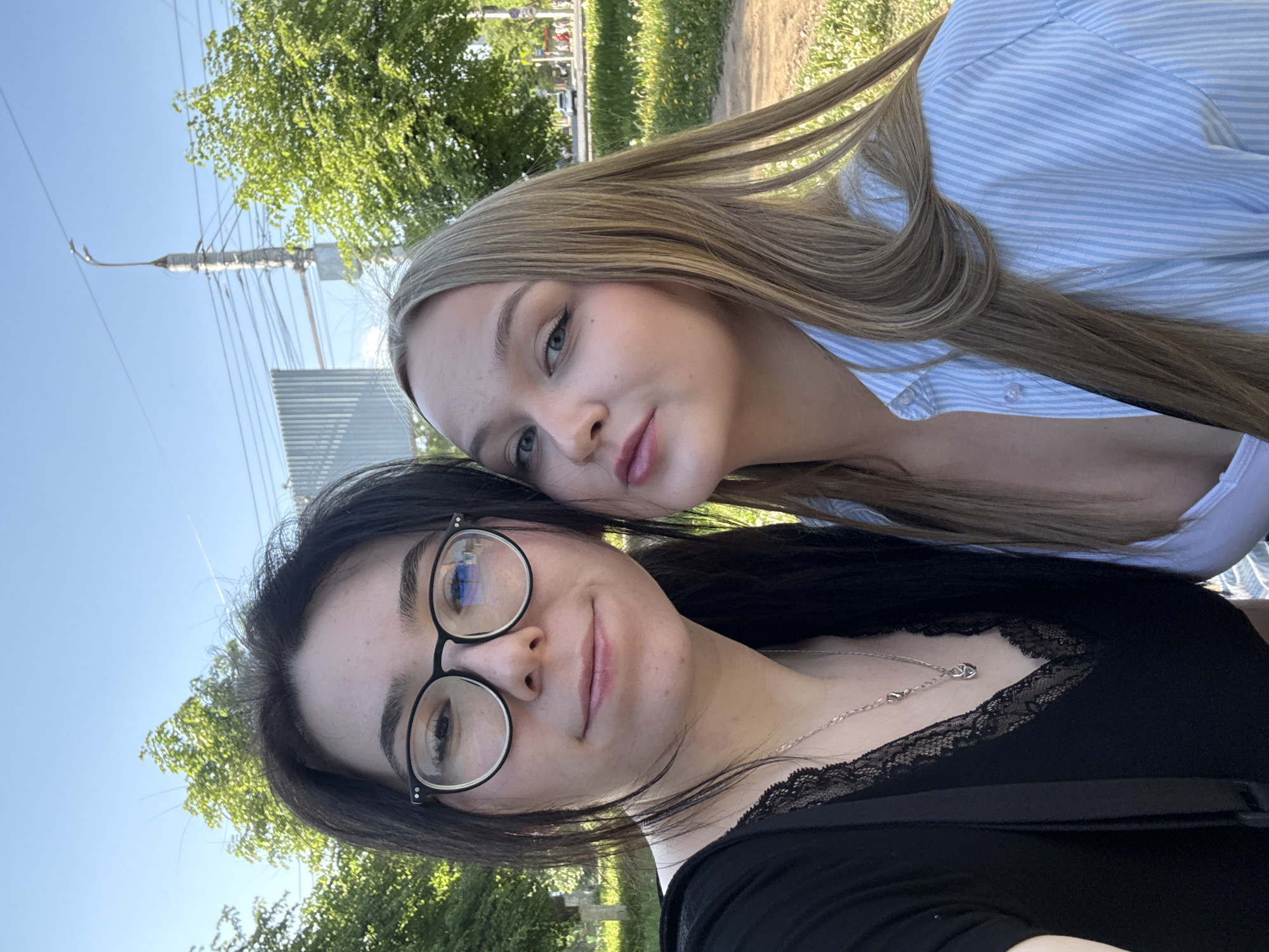 Two young women are posing for a selfie outdoors, possibly in a park or green area. The woman in the foreground has dark hair and is wearing glasses and a black top with lace detailing at the neckline. She is also wearing a silver necklace. The woman behind her has long, blonde hair and is wearing a light blue and white striped shirt. The background includes trees, grass, a utility pole with wires, and a partial view of a building in the distance. The lighting suggests it is daytime. The city location is Izhevsk, Russia.