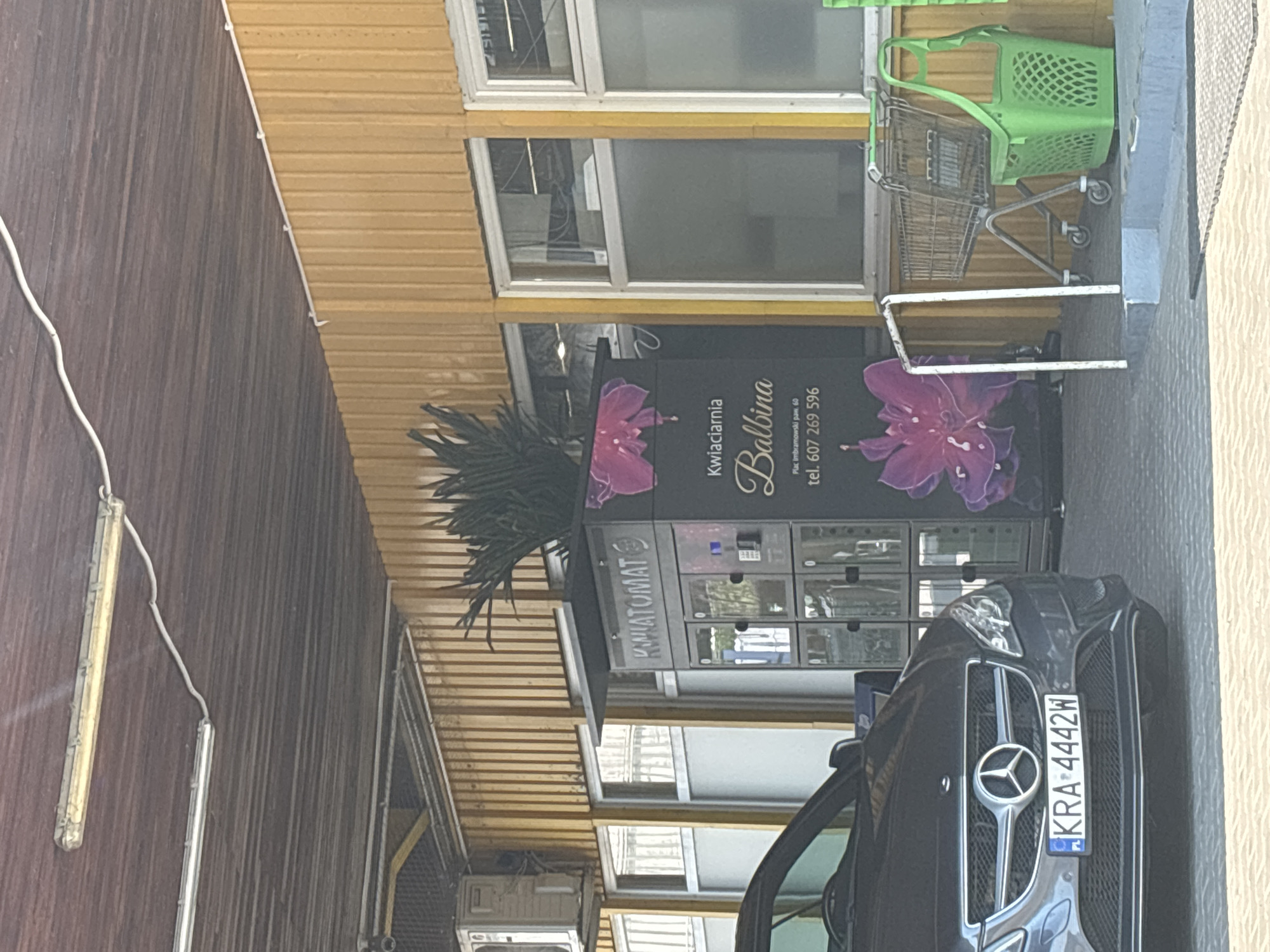 This image captures a street-level view of a flower vending machine, identified by the Polish word "KWiatomat," located in Krakow, Poland. The vending machine is dark in color with graphical elements depicting large, stylized pink flowers. Text on the machine indicates it is for "Kwiaciarnia Balbina," a flower shop, and provides a Polish phone number, 607 269 596, and an address, "Plac Imbramowski paw. 60."
To the left of the vending machine, the front portion of a dark grey Mercedes-Benz car is visible, with a Polish license plate reading "KRA 4442W." The car is parked on a paved surface. To the right of the vending machine, a metal shopping cart and a bright green plastic basket are positioned.
The background consists of a yellow, corrugated metal wall and a wooden ceiling structure with visible fluorescent lighting fixtures. Windows with frosted glass are present on the yellow wall. An air conditioning unit is mounted on the building structure above the windows on the left. The overall scene appears to be an outdoor service area or a covered parking space.