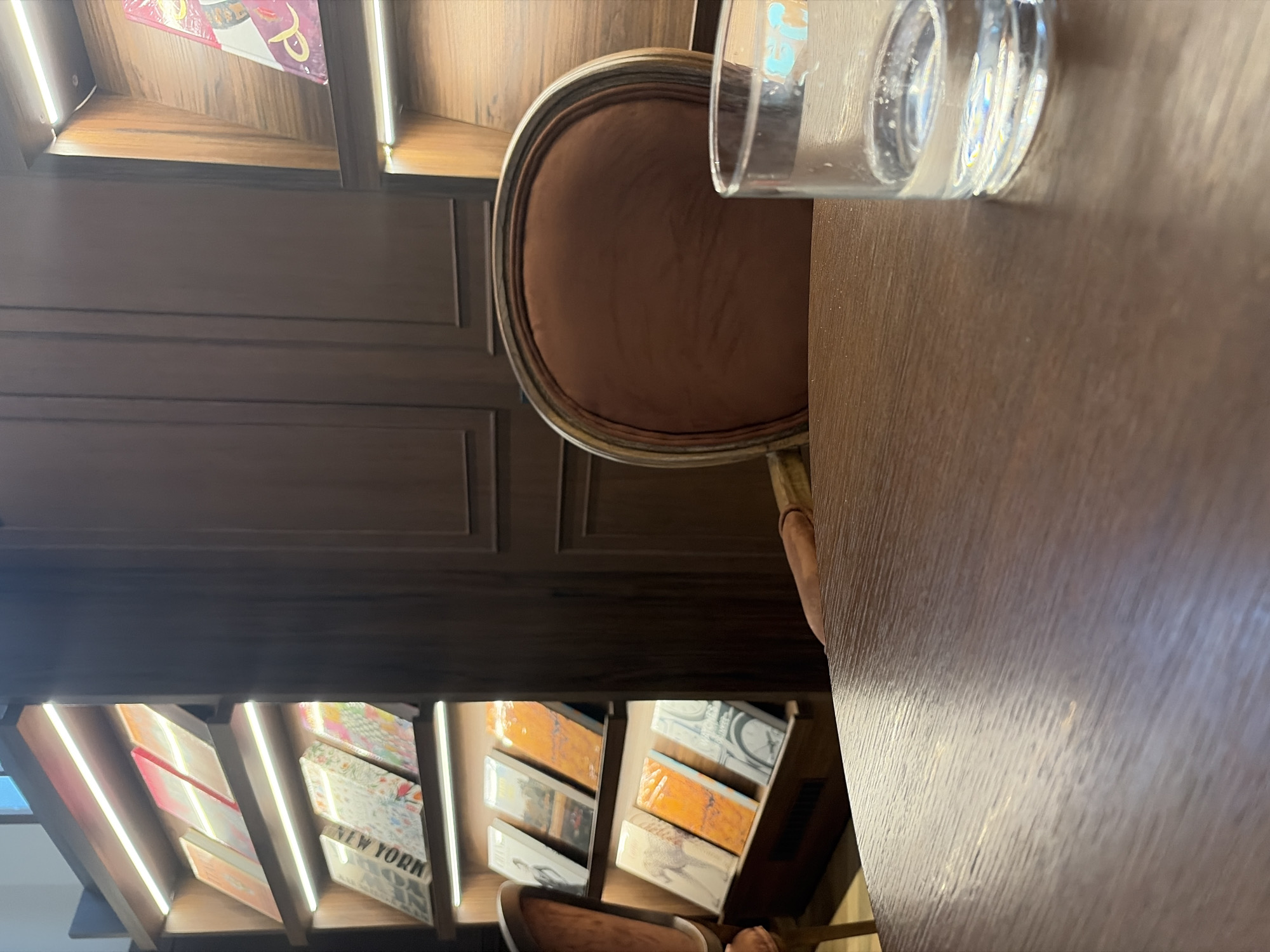 This is a photograph of a cozy corner in a library or cafe located in Minsk, Belarus. The scene is dominated by dark wood paneling and shelving filled with books. In the foreground, a portion of a dark wood table is visible, with a glass of water placed on it to the right. Behind the table, a plush, upholstered armchair with a dark brown fabric and a curved wooden frame is positioned. Further back, shelves are illuminated by soft strip lighting, displaying a variety of books with colorful covers. The overall atmosphere suggests a quiet and inviting space for reading or relaxation.