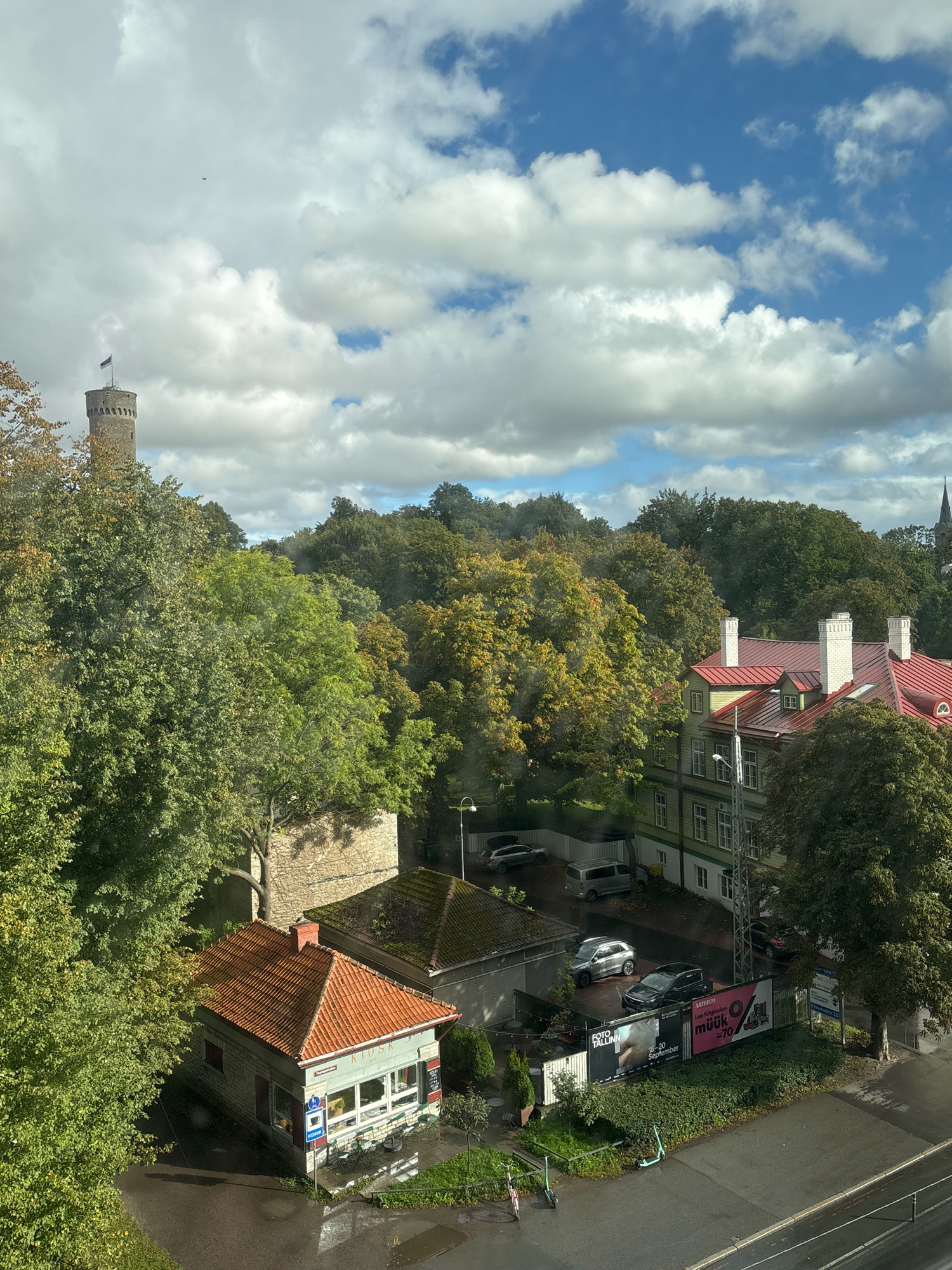 The image depicts a view from a window in Tallinn, Estonia, overlooking a street scene with trees and buildings under a cloudy sky. In the background, a prominent circular stone tower with a black flag atop it stands against the sky, likely part of the city's historical fortifications. A cluster of mature trees, displaying a mix of green and autumnal yellow foliage, dominates the mid-ground.
In the foreground, a small, single-story building with a red-tiled roof, identified by the sign "KIOSK", is visible. Adjacent to the kiosk, a paved area shows parked vehicles, including a silver SUV, a black car, and a grey van. A billboard with text including "FOTO TALLINN" and event dates is also present. Further to the right, a two-story building with a red-tiled roof and green siding, featuring white trim and multiple chimneys, is partially visible. The street in front of the buildings appears wet, suggesting recent rain. A lamppost is situated to the right of the buildings, and two kick scooters are parked on the side of the road. The overall atmosphere is urban, with elements of nature and history.