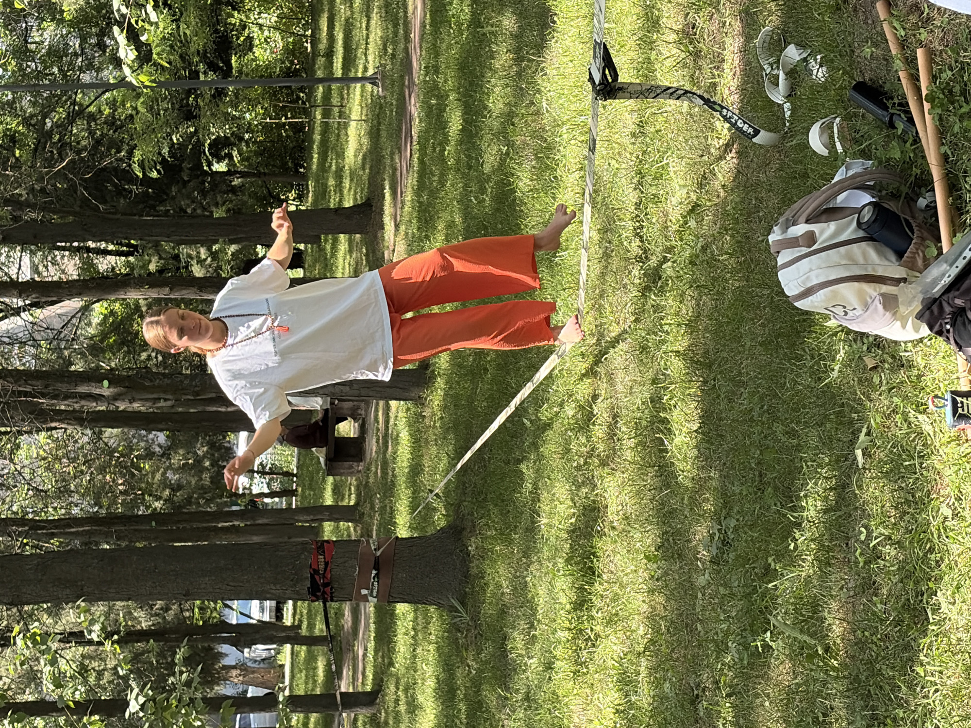 A young person with blonde hair pulled back is slacklining in a park. They are wearing a white t-shirt and orange pants. The slackline is strung between two trees. The person is balanced on the slackline with their arms extended for stability. The ground is covered in green grass and dappled with sunlight. In the foreground, a backpack, a water bottle, and some wooden sticks are visible on the grass. Other trees and foliage surround the area, and a wooden bench can be seen in the background. The scene appears to be a recreational activity taking place outdoors in a city park.