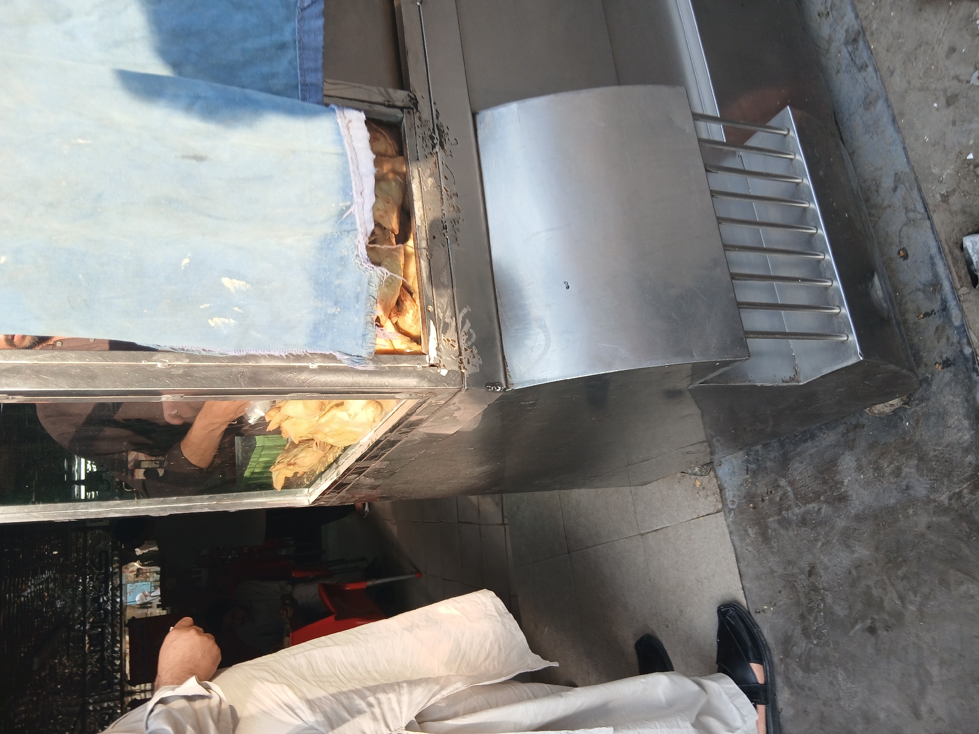 The image depicts an outdoor food stall in Peshawar City Tehsil, Pakistan. A person wearing a light-colored shalwar kameez and black sandals is visible on the left, facing away from the camera. To the right, a stainless steel food display case contains several fried food items, possibly samosas, illuminated from within. A blue cloth is draped over the top of the stall. The ground is a mix of concrete and tiled flooring. In the background, hints of a dimly lit interior with seating and other people suggest a bustling environment. The stall appears to be situated in a market or street setting.