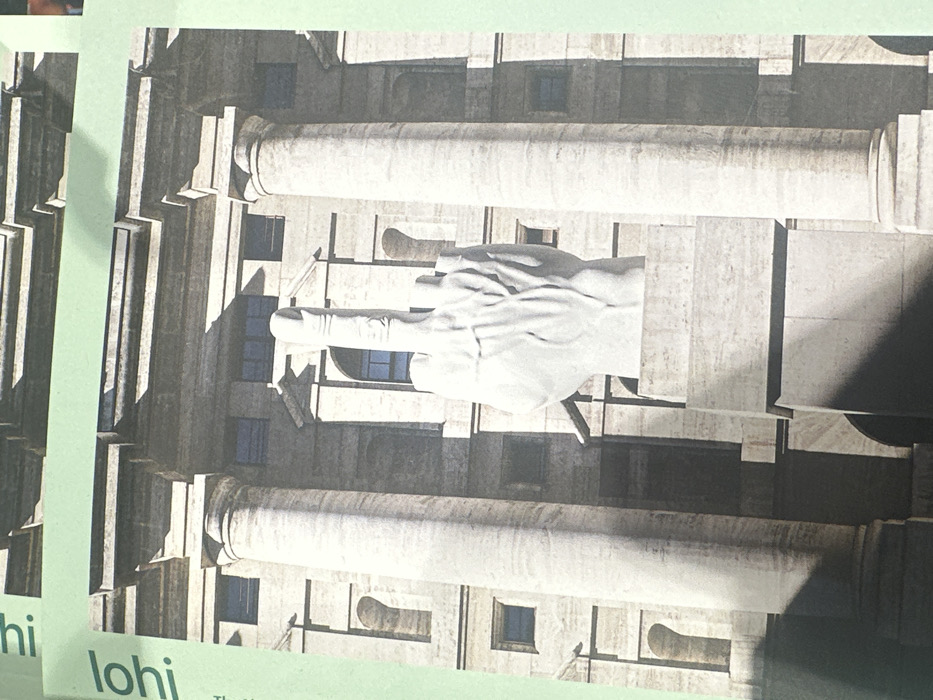 The image displays a photograph of a large, white marble sculpture of a human left hand with all fingers except the middle finger curled downwards, presenting an upward-pointing middle finger. This artwork, known as "L.O.V.E." by Maurizio Cattelan, is situated on a multi-tiered stone pedestal. Behind the sculpture stands a large, neoclassical building constructed of light-colored stone, featuring several prominent fluted columns with classical capitals, multiple rectangular window openings, and decorative architectural elements. The scene is illuminated by direct sunlight, creating distinct shadows. The depicted location is Piazza Affari in Milan, Italy, specifically in front of the Palazzo Mezzanotte, which houses the Borsa Italiana (Italian Stock Exchange). The photograph itself appears to be of a printed page, evidenced by a light green border and partial text "lohi" visible along the left and top edges.