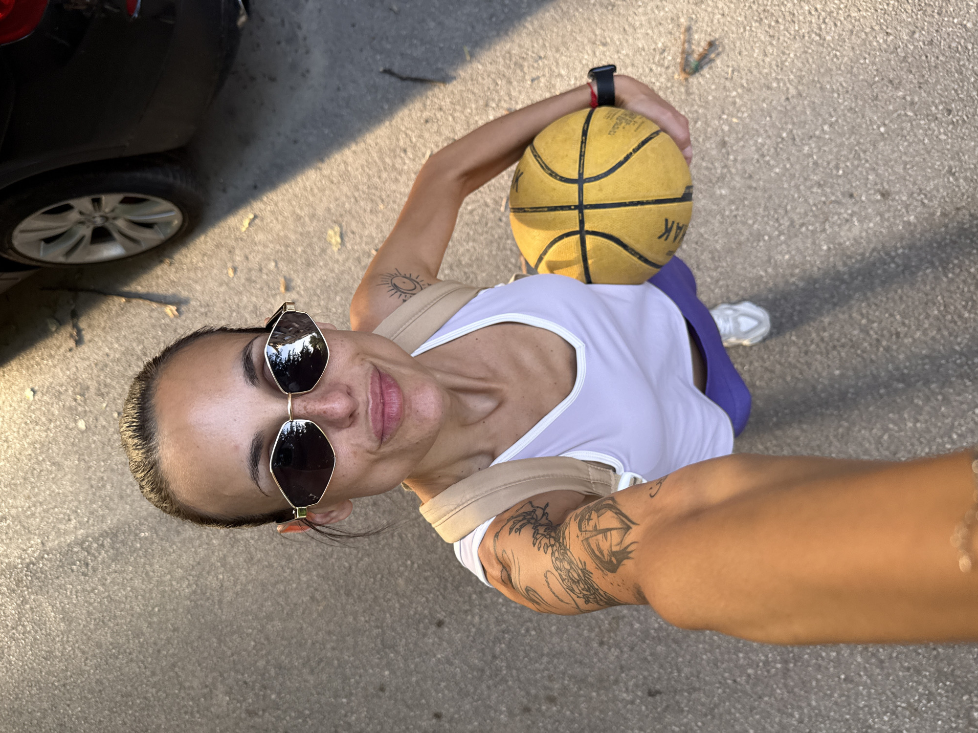 A high-angle selfie shows a woman holding a yellow basketball. She wears sunglasses and a white tank top. A black car is visible in the background, parked on a paved surface. The location is potentially in the city of Pleven, Bulgaria; this is speculative and based solely on the user's instructions. There are tattoos on her left shoulder and forearm. She wears a wristwatch on her left wrist. The background surface appears to be asphalt or concrete. A white shoe is visible near the bottom right.
