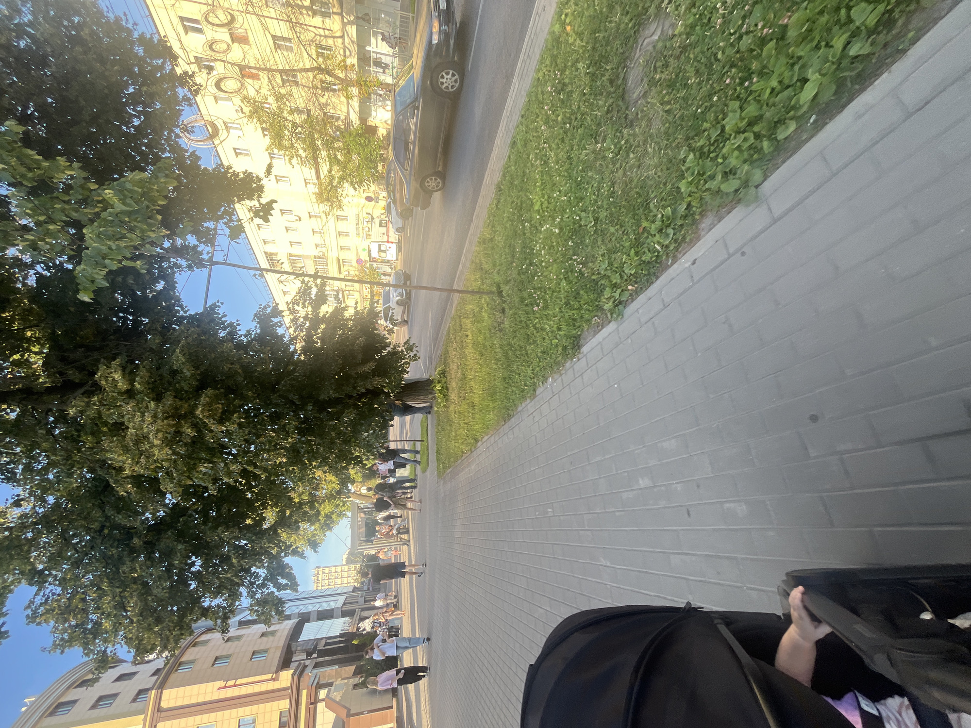 The image depicts a street scene in Novaya Usman, Russia, on a sunny day. The perspective is from the viewpoint of someone pushing a stroller. In the foreground, a black stroller is visible. The scene is a paved sidewalk with a strip of green grass and trees on the left. Across the street, there are several buildings, including a light-colored apartment building with multiple windows and some decorative elements. Further down the street, cars are parked and moving.
Several people are scattered along the sidewalk, walking and standing. Some are dressed casually, suggesting a leisurely outing. The sunlight creates bright highlights and shadows, indicating it's likely late afternoon or early morning. The clear blue sky suggests good weather. The overall mood appears relaxed and everyday. No discernible text is visible in the image.