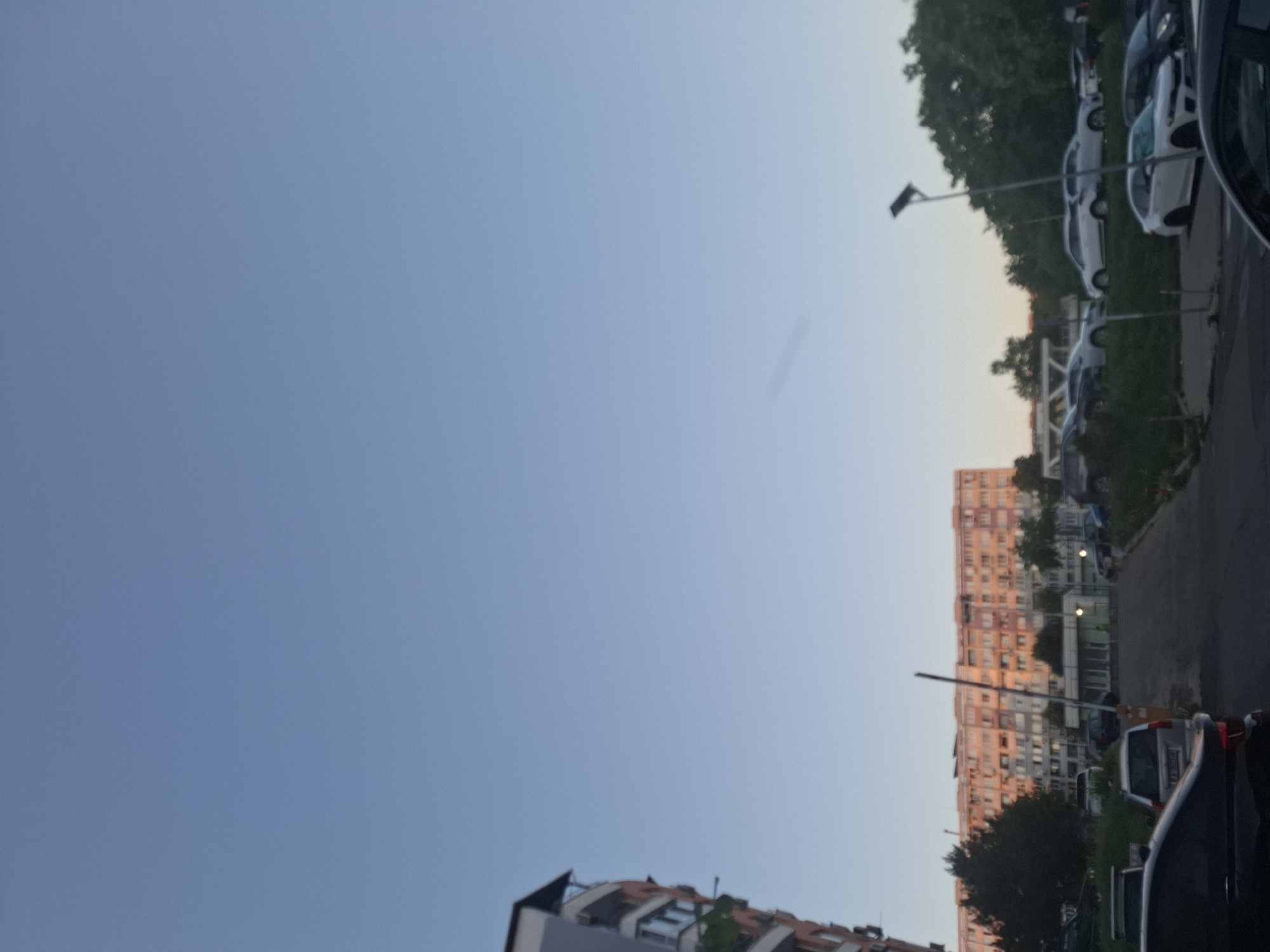An outdoor, low-angle, vertical, full shot shows a clear sky over a residential area. The sky is a pale blue with a faint, wispy cloud. In the lower half of the image, a large, pink-bricked, apartment building with many windows and balconies dominates the left side. To the right of the building, a parking lot filled with various cars is visible, with green grass and trees bordering the area. A street runs through the center of the image, with a white pole and a barrier gate on the left side. The scene appears to be at dusk, with the sun casting a warm glow on the apartment building.