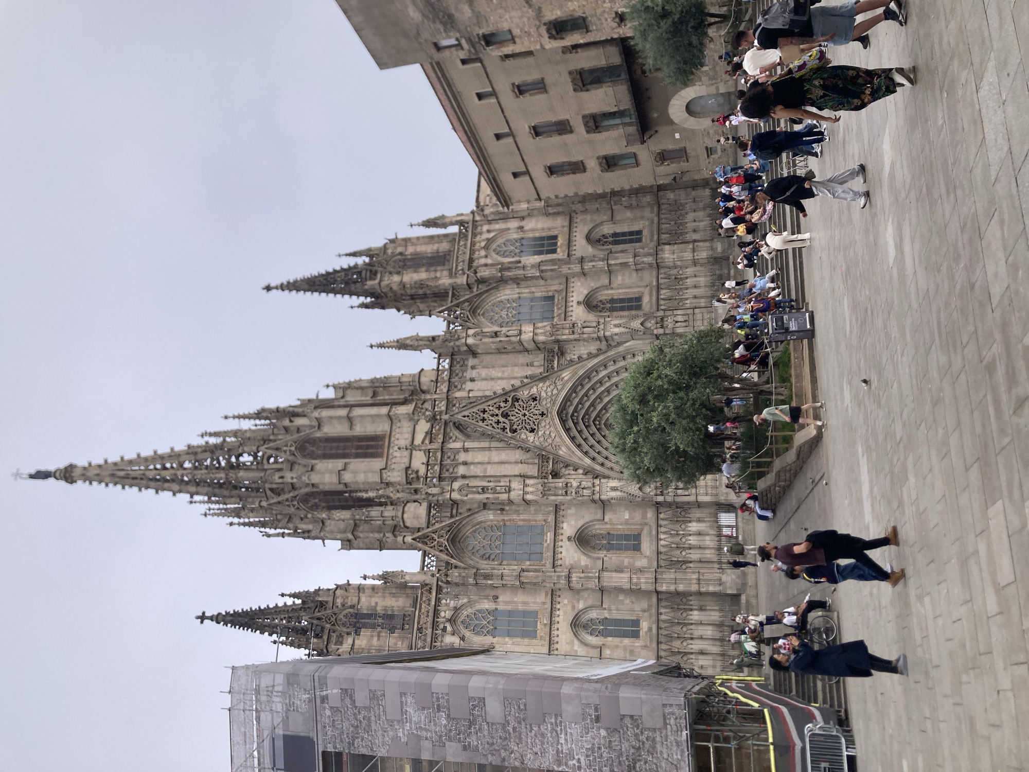 The image shows the Gothic Barcelona Cathedral in Barcelona, Spain. The cathedral's facade is visible with its intricate carvings, spires, and windows. Parts of the cathedral are covered in scaffolding. In front of the cathedral, there is a paved square filled with people. Some people are sitting on steps. Several people are walking around the square. A building is also seen on the edge of the square. The sky is overcast.