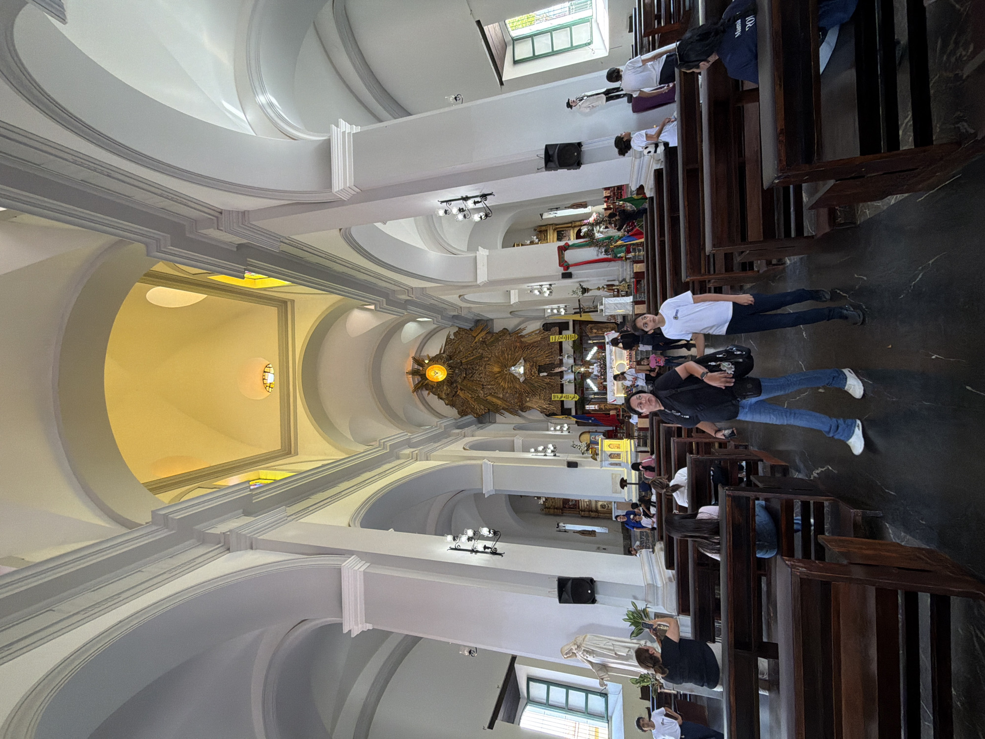 The image depicts the interior of the Metropolitan Cathedral of Ciudad Bolivar, Venezuela. The cathedral features a high, arched ceiling painted in white and yellow hues. Ornate gold decorations are prominently displayed behind the altar. Pews are arranged in rows along the sides of the nave. Several people are present; some are seated in the pews, while two individuals stand in the aisle. One person appears to be taking a photograph with a mobile phone. Windows can be seen along the sides of the cathedral. Speakers are mounted on the walls.