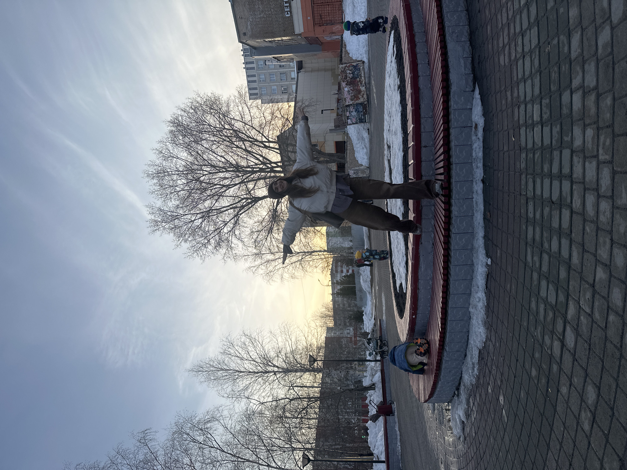 A young woman with long brown hair stands on a circular, elevated seating area in Surgut, Russia, with her arms outstretched. She wears a light-colored jacket, brown pants, and a dark hat. A light-colored purse hangs from her shoulder. The seating area is constructed of red and gray wood and has a concrete border. Patches of snow are visible around the perimeter of the seating area and on the adjacent walkway, which is paved with gray blocks. In the background are bare trees, buildings, and a section of sky. A child wearing a blue coat and colorful hat sits on the seating area near the bottom of the frame. Another child is visible in the upper right corner, dressed in black and wearing a green hat.