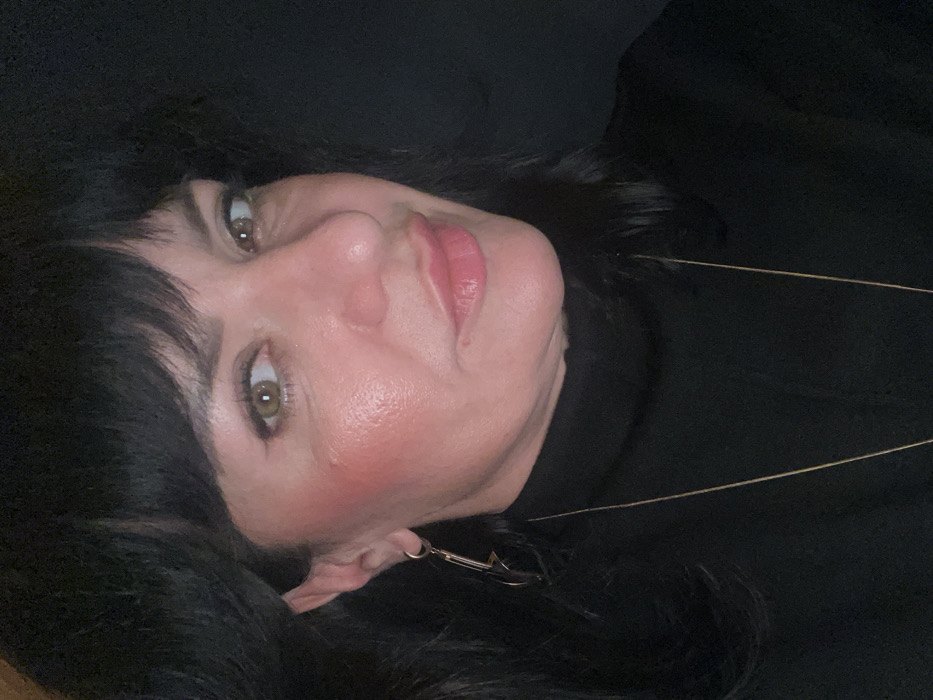 A woman with black hair and fair skin is lying down, her head tilted to the side. She has green eyes, pink lipstick, and blush on her cheeks. She's wearing a black turtleneck sweater and a thin gold necklace. An earring is visible on her left ear. The background is dark and blurry. The image appears to be taken at a close-up angle.