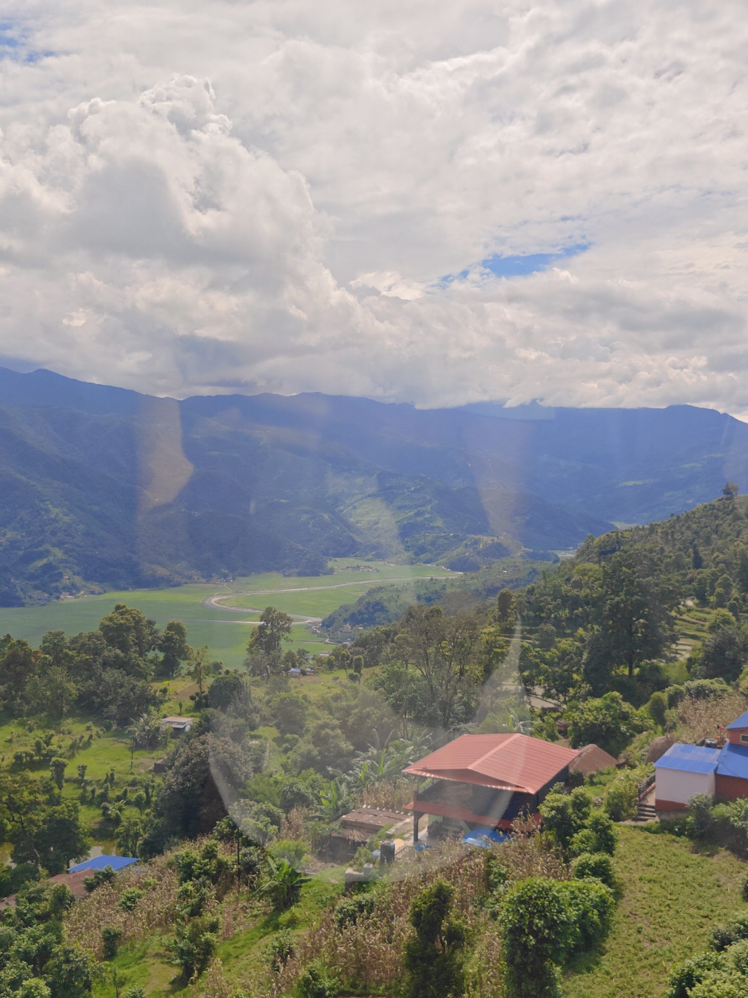 The image captures a panoramic view of Pokhara, Nepal, showcasing the majestic Annapurna mountain range in the background, partially obscured by dramatic clouds. Sunbeams pierce through the clouds, casting ethereal rays onto the verdant landscape. In the mid-ground, a serene lake, likely Phewa Lake, is visible, its tranquil waters reflecting the surrounding greenery. The foreground is dominated by rolling hills covered in dense foliage and agricultural terraces. Several small houses and buildings are scattered across the landscape, including a prominent building with a red tiled roof and an attached structure with a thatched roof. The overall scene conveys a sense of natural beauty and rural tranquility characteristic of Pokhara.