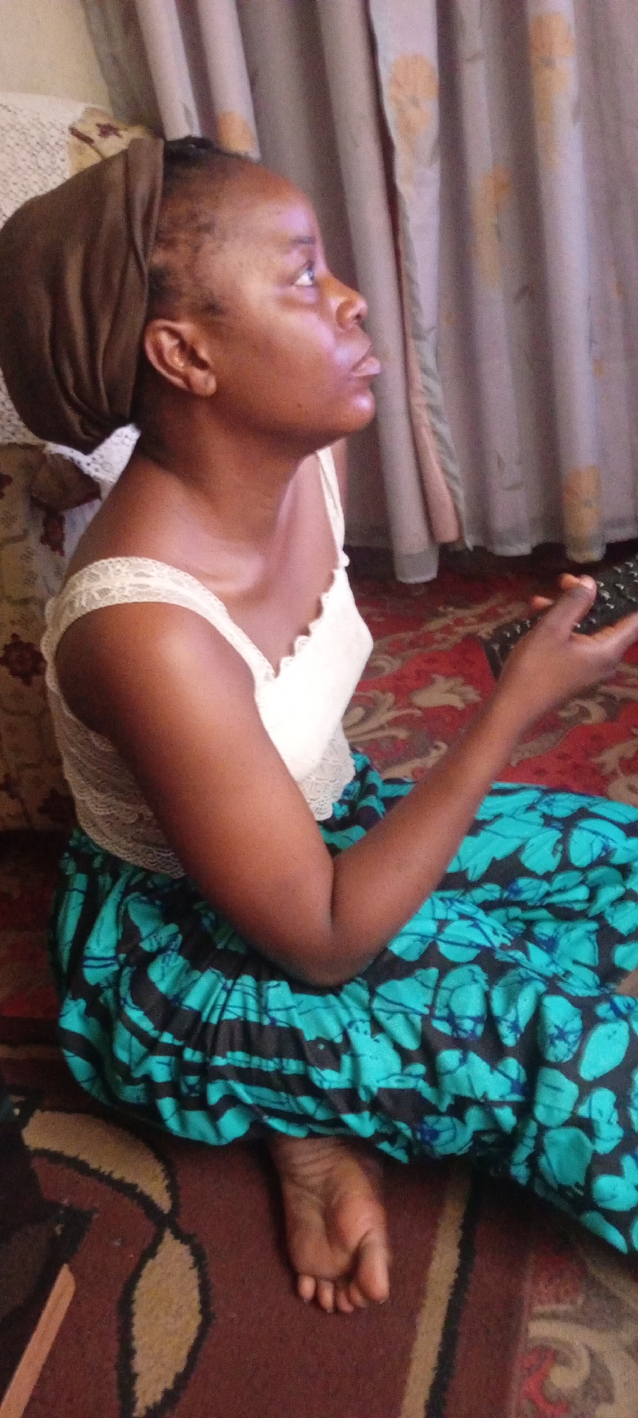 A dark-skinned woman sits on a patterned rug in a room, her gaze directed upwards and to the right. She is wearing a light-colored, possibly white, sleeveless top with lace detailing, and a vibrant, turquoise and black patterned skirt. A brown headscarf covers her hair. She holds a black remote control in her right hand. The background includes patterned curtains and a portion of a sofa or cushion. The location is Gawuna, Nigeria.