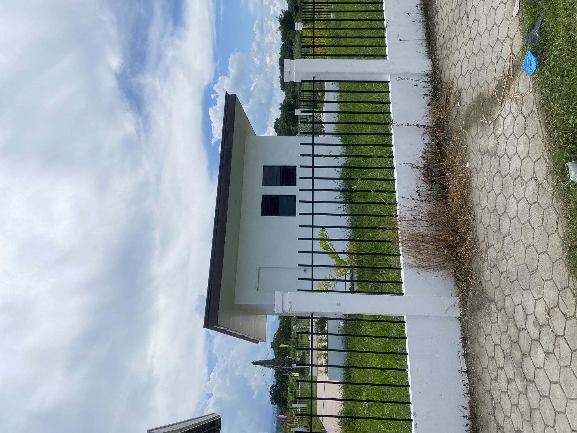 The image shows a small, white, rectangular building with a brown, sloped roof, situated behind a white concrete wall and a black metal fence. The building has two windows with dark frames and a white door on the left side. The fence encloses a grassy area with some scattered dry vegetation. In the background, there are trees, hills, and a cloudy sky with patches of blue. The foreground features a hexagonal paver walkway and grass. A blue plastic bag is visible on the grass near the walkway. The scene appears to be an outdoor construction site or a newly developed area in Davao City, Philippines, given the tropical vegetation and the style of the building. There are no people visible in the image.