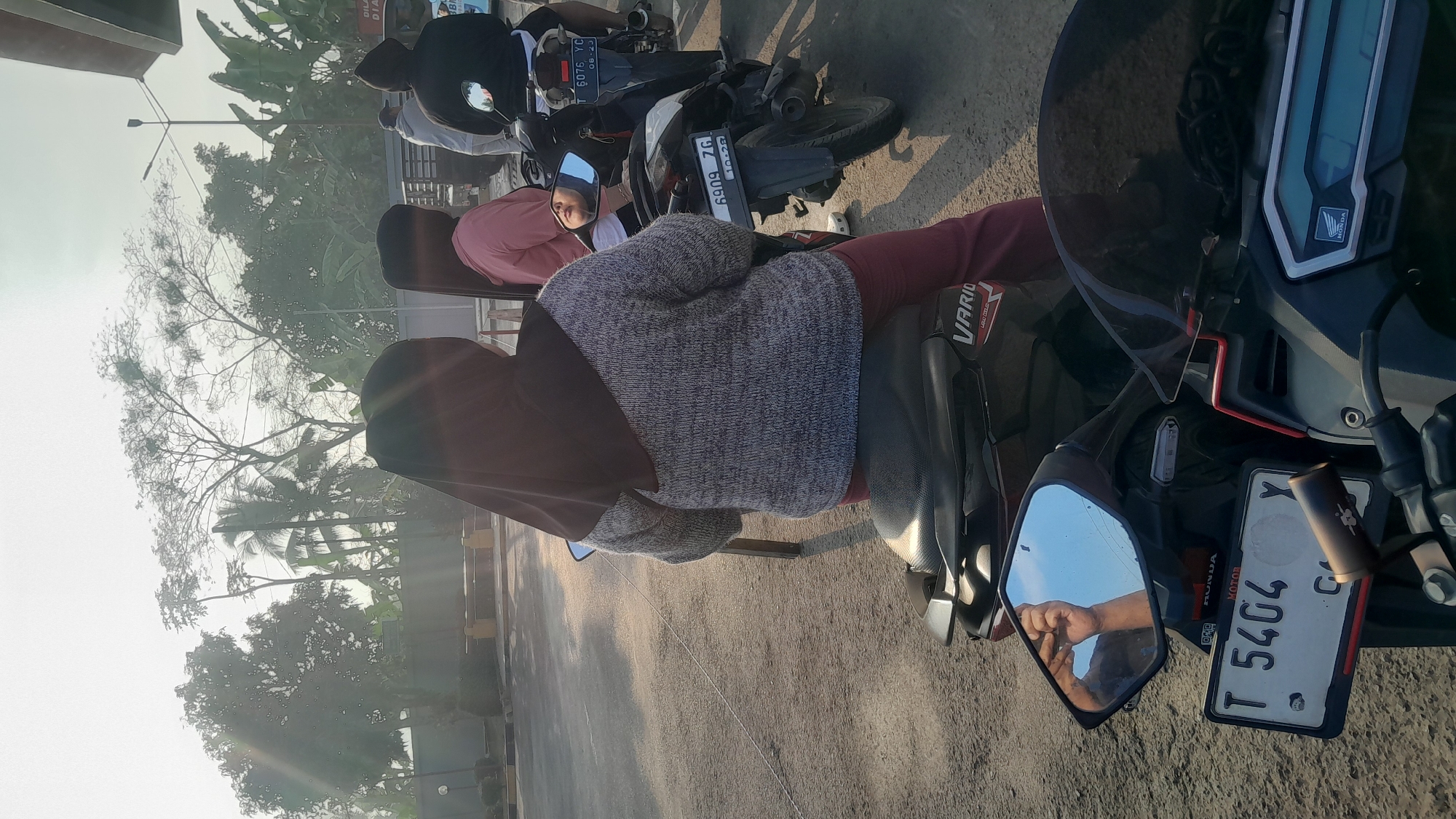 In Subang, Indonesia, on a sunlit day, two women and two men are on motorcycles. The two women are wearing hijabs, one in a dark brown and the other in a lighter shade. The first woman is in the foreground, facing away from the camera, wearing a grey knitted sweater and maroon pants. The second woman, next to her, is wearing a pink top. Two men are behind them on motorcycles. The environment appears to be an outdoor area, possibly a parking lot or road, with trees and a clear sky visible. The motorcycles are black and have license plates. The license plate of the motorcycle in the foreground reads "T 5404 Y". The license plate of the motorcycle behind the first woman reads "6909 ZG". The license plate of the motorcycle behind the second woman reads "T 6076 YC". The scene is brightly lit by the sun, casting shadows.