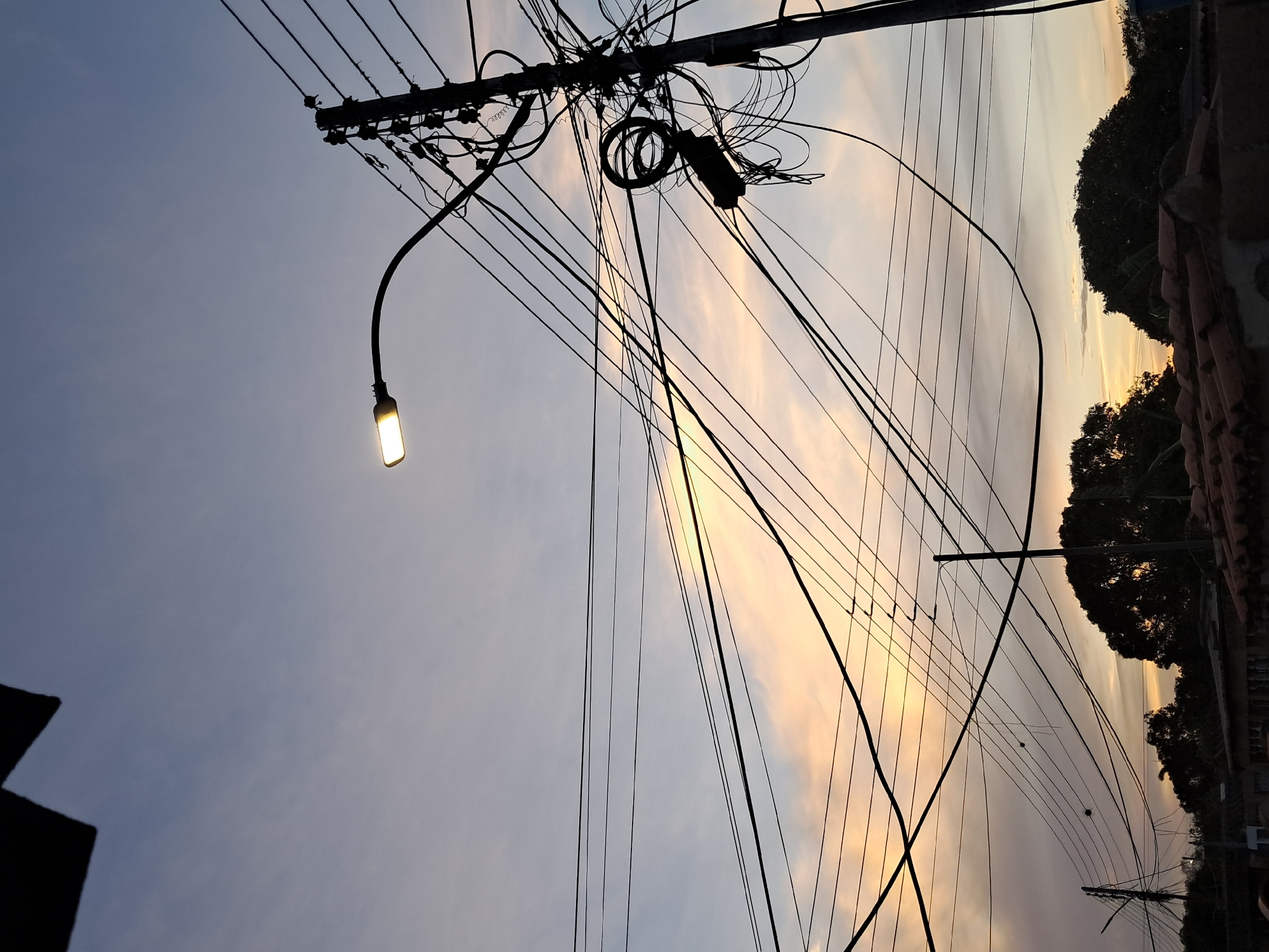 The image captures a scene in Juan Griego, Venezuela, likely at dusk or dawn, as suggested by the soft orange and gray sky. A utility pole with a streetlight is the focal point. The pole is cluttered with numerous electrical wires and cables, indicative of a dense network. The streetlight is illuminated. Silhouetted trees and a building can be seen against the sky, adding depth to the composition. The photo is taken from a low angle, looking upward toward the sky and infrastructure.