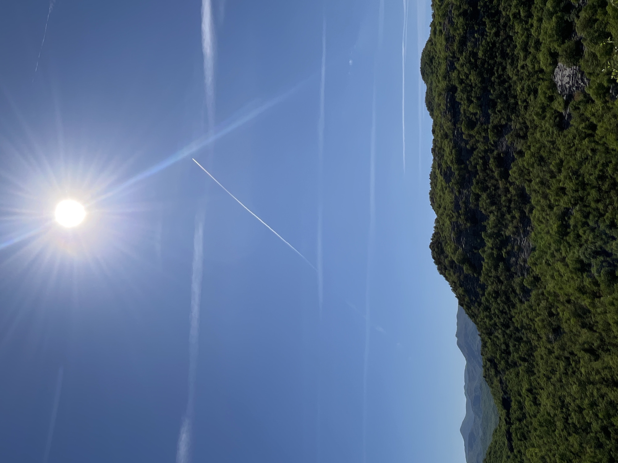 The image shows a daytime landscape in Gornja Brova, Montenegro. A clear blue sky occupies the upper left portion of the frame, with a bright sun emitting rays. Several contrails are visible across the sky, some intersecting. The right side of the image features a densely forested mountainside, showcasing varying shades of green. Lower in the frame, the peaks of other mountains are visible.