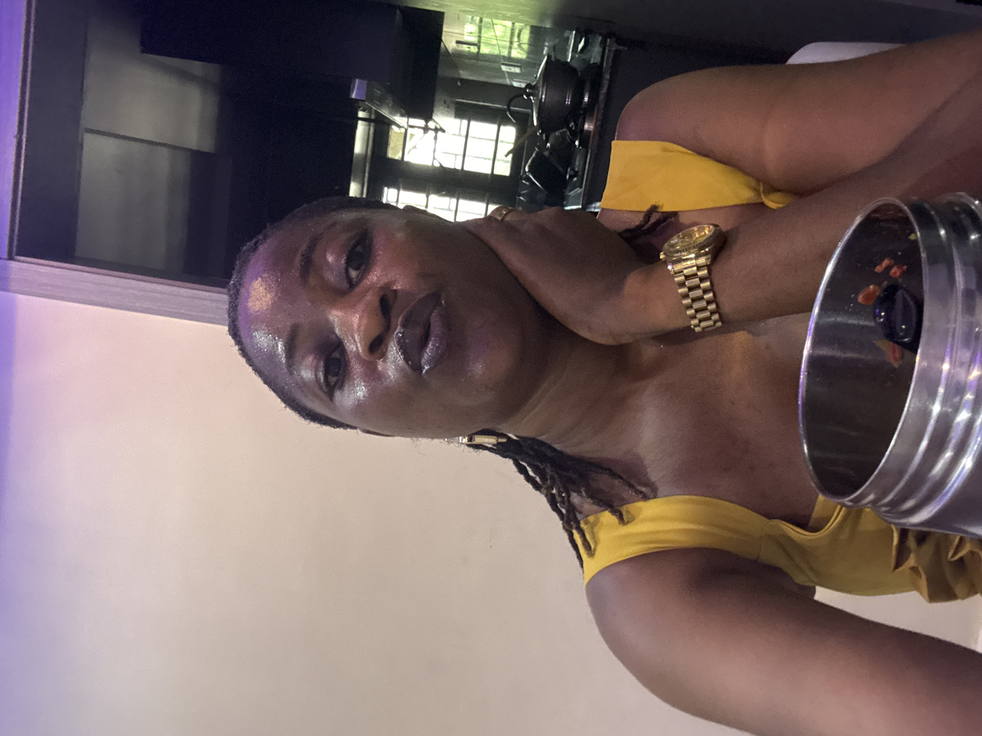 A woman is the primary subject of the image. She is wearing a yellow top and a gold watch. Her skin appears to be glistening, possibly due to sweat or moisture. She has dark hair, braided with some gold accessories. A metal container, seemingly empty, is positioned in the lower right foreground. The background shows a partially visible window with a grid pattern and an indoor setting with some equipment that might be related to a kitchen. The context suggests the photo may have been taken indoors, perhaps in a restaurant or home. There is no direct indication that the image was taken in the city of Ajara, Nigeria.