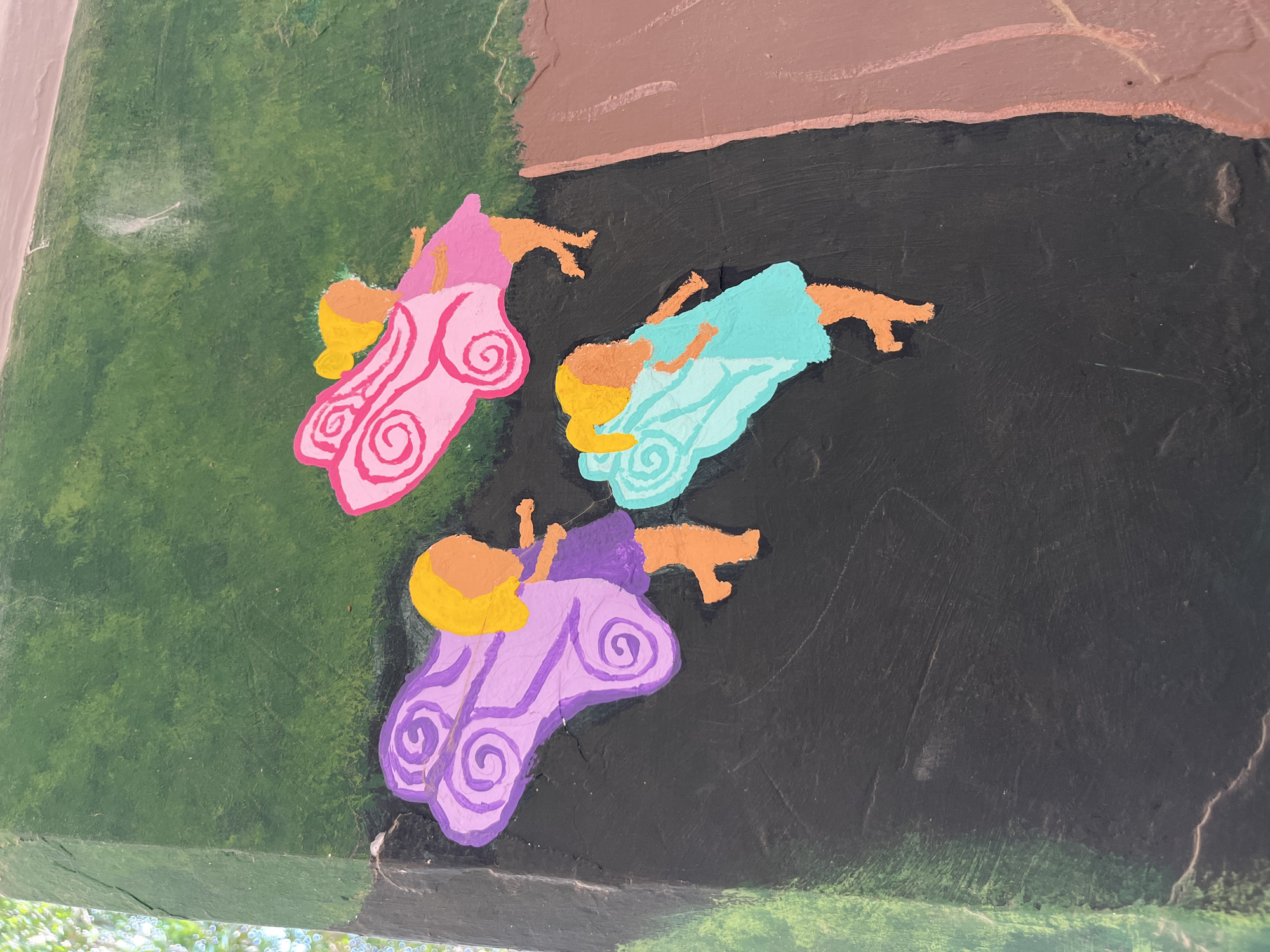 The image depicts a painted mural of three figures lying down, possibly fairies, on a rough, textured surface that appears to be part of a larger structure or wall. The figures are rendered in a simplified, cartoonish style with distinct outlines and flat colors.
Each figure has tan skin and golden-yellow hair. They are wearing differently colored dresses: one pink, one light blue, and one purple, all adorned with a spiral pattern on the skirt. The figures are positioned close to each other and appear to be on a dark, possibly black, background.
The surrounding environment includes sections of green and brown paint, suggesting a depiction of foliage or landscape. The image is taken at an angle, possibly from below, as evidenced by a glimpse of a bright, blurred area at the bottom, potentially sky or sunlight filtering through trees.