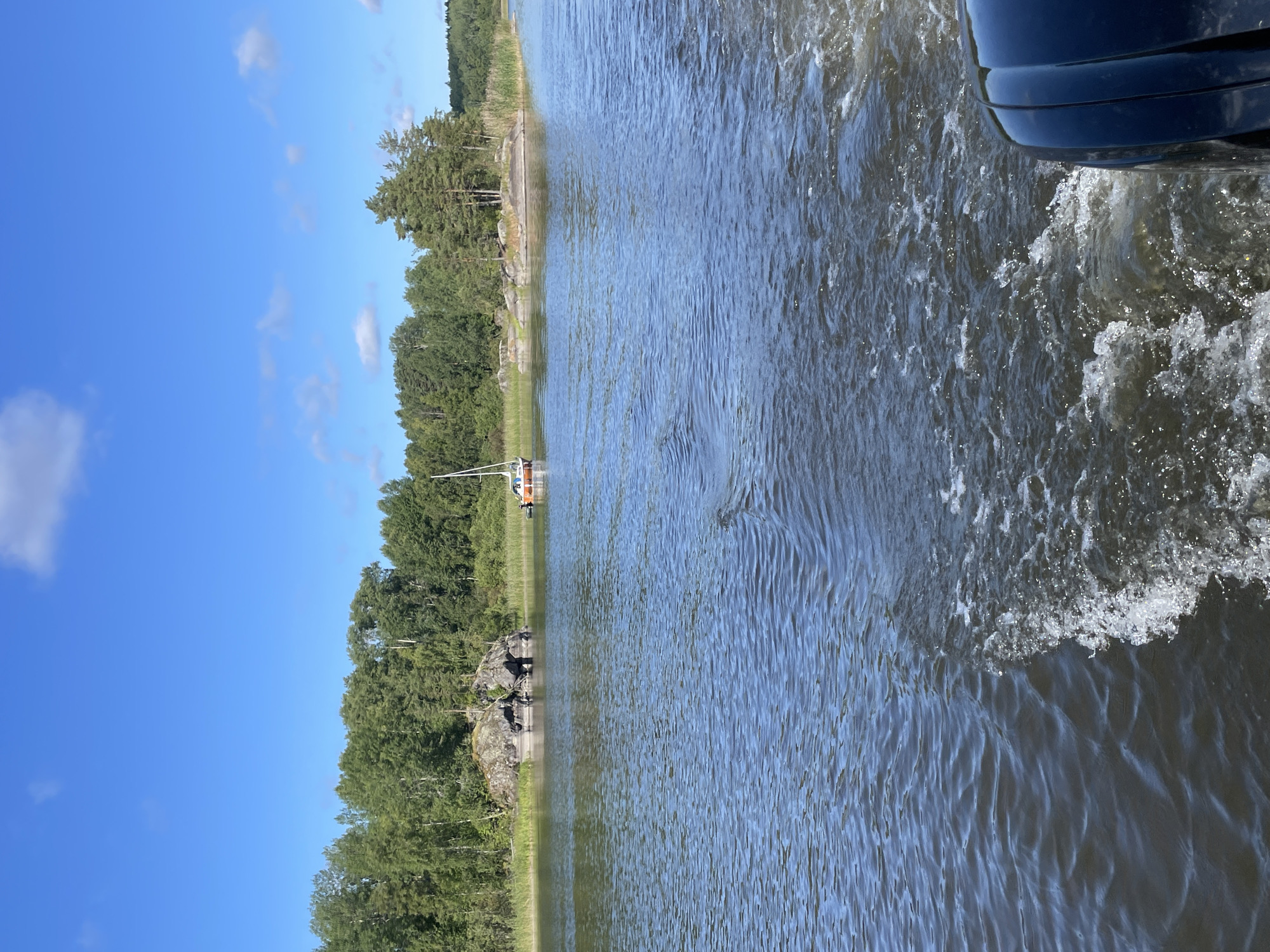 A wide-angle, eye-level shot captures a tranquil water scene under a bright blue sky with scattered white clouds. In the foreground, dark blue water ripples, disturbed by the wake of an unseen boat, part of which is visible on the right side of the frame, appearing as a black outboard motor. In the middle ground, a small sailboat with an orange hull and a white sail navigates the calm water. Beyond the sailboat, a rocky outcrop covered in greenery juts out of the water, backed by a dense forest that stretches across the horizon. The trees are a lush green, with a few taller pine trees interspersed. The scene is serene and natural. The image is not set in the city of Berozovo, Russia; it depicts a natural body of water with a boat and forested shoreline.