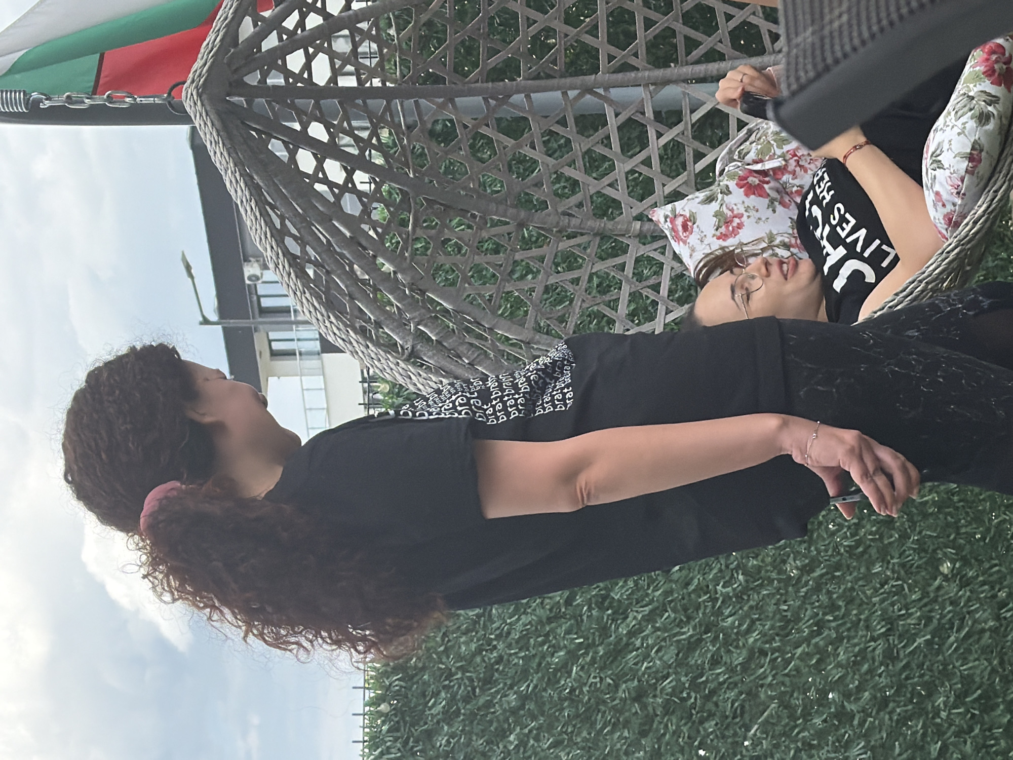 In Sofia, Bulgaria, an outdoor scene depicts two individuals. On the left, a person with long, curly dark hair tied back with a pink scrunchie stands, wearing a black t-shirt and dark pants. They are facing to the right and holding a dark-colored smartphone in their right hand. To the right, another person, wearing glasses and a black t-shirt with white text reading "JACK LIVES HERE," is relaxing in a wicker hanging chair. They are looking at a smartphone held in their right hand, resting on floral-patterned cushions. A Bulgarian flag is visible in the background, attached to a pole. The environment includes a green hedge, a modern building, and a street lamp.