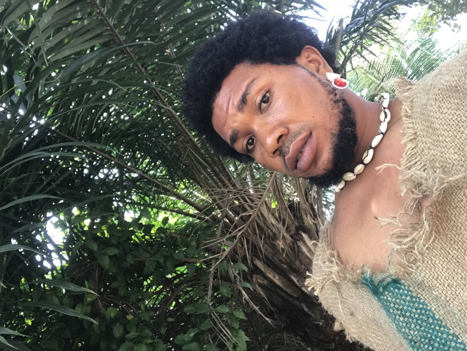 A low-angle portrait depicts a single individual, appearing male, with dark curly hair and a trimmed beard, gazing directly at the camera. The subject wears a rustic, burlap-like garment with frayed edges and a turquoise fabric stripe on the left shoulder. A necklace composed of multiple cowrie shells is worn around the neck, and a large earring featuring a prominent red center stone surrounded by white accents is visible on the right ear. The background consists of dense, verdant foliage, including distinct palm fronds and other green leaves, illuminated by natural sunlight filtering through the canopy, indicative of an outdoor, tropical setting within the city of Igbologun, Nigeria.