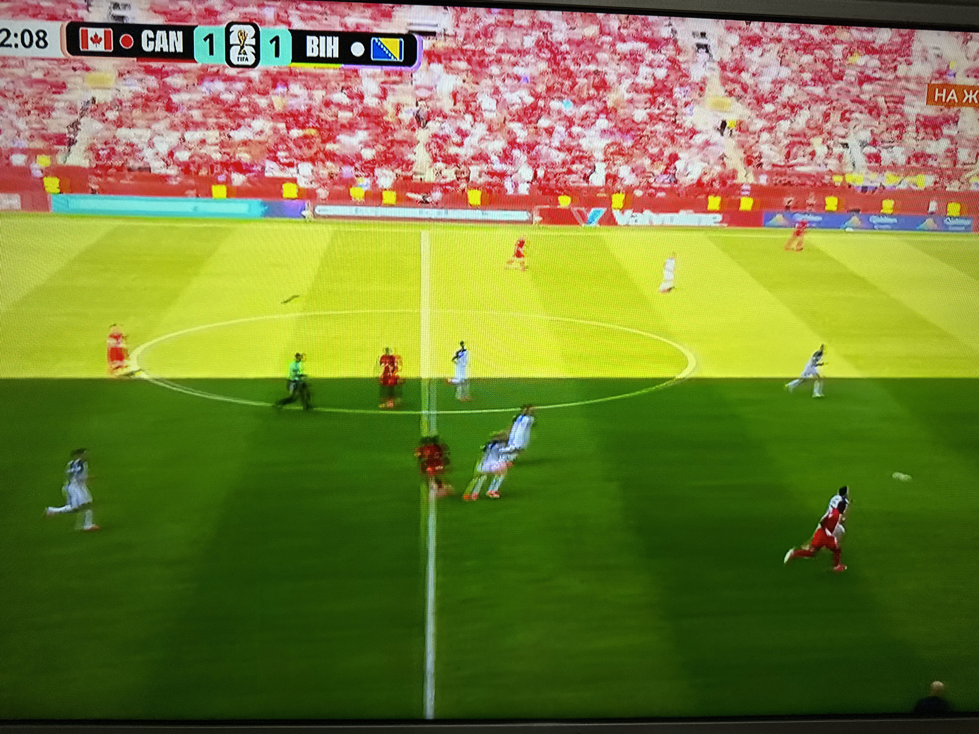 The image is a screenshot from a soccer match being broadcast on television. In the top left corner, a timer displays "2:08". To the right of the timer, a scoreboard indicates the teams "CAN" and "BIH" are tied 1-1. The background shows a stadium filled with a crowd dressed primarily in red. The field is green with yellow markings. Several players in white and red uniforms are visible on the field, some in motion as if participating in the game. A portion of a sign in Cyrillic script is visible on the right side of the screen. The photograph was taken of a television screen, as evidenced by the pixelation and glare. The provided location of Targovishte, Bulgaria is not supported by any visual information in the image.