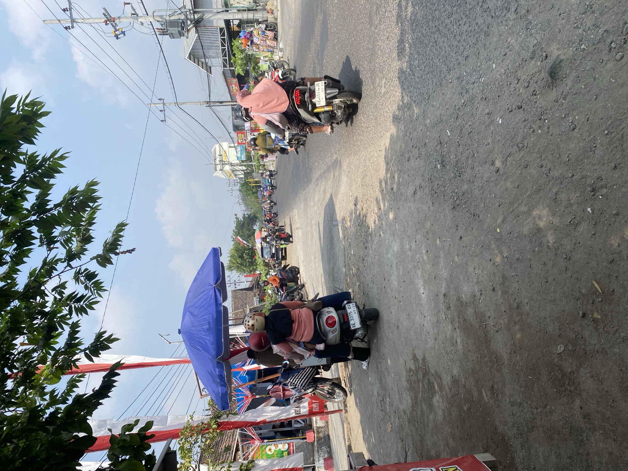 The image depicts a sunny daytime scene on a street in Sempol, Indonesia. In the foreground, a dirt and gravel shoulder runs alongside a paved road. A blue umbrella is set up on the side of the road, providing shade for several people on a scooter. The scooter carries at least three individuals, including two adults and a child, all wearing helmets. The adults appear to be a woman and a man, with the woman holding the child. The scooter has a license plate visible. Behind the umbrella, the street is busy with traffic, predominantly motorcycles, some carrying multiple passengers. Buildings line the right side of the street, with shops or stalls visible. Red and white banners or flags are displayed on the left side of the road. The sky is clear blue with some scattered clouds. Trees with green leaves are visible in the upper left corner, casting shadows on the ground. The overall impression is a typical street scene in a populated area of Indonesia.