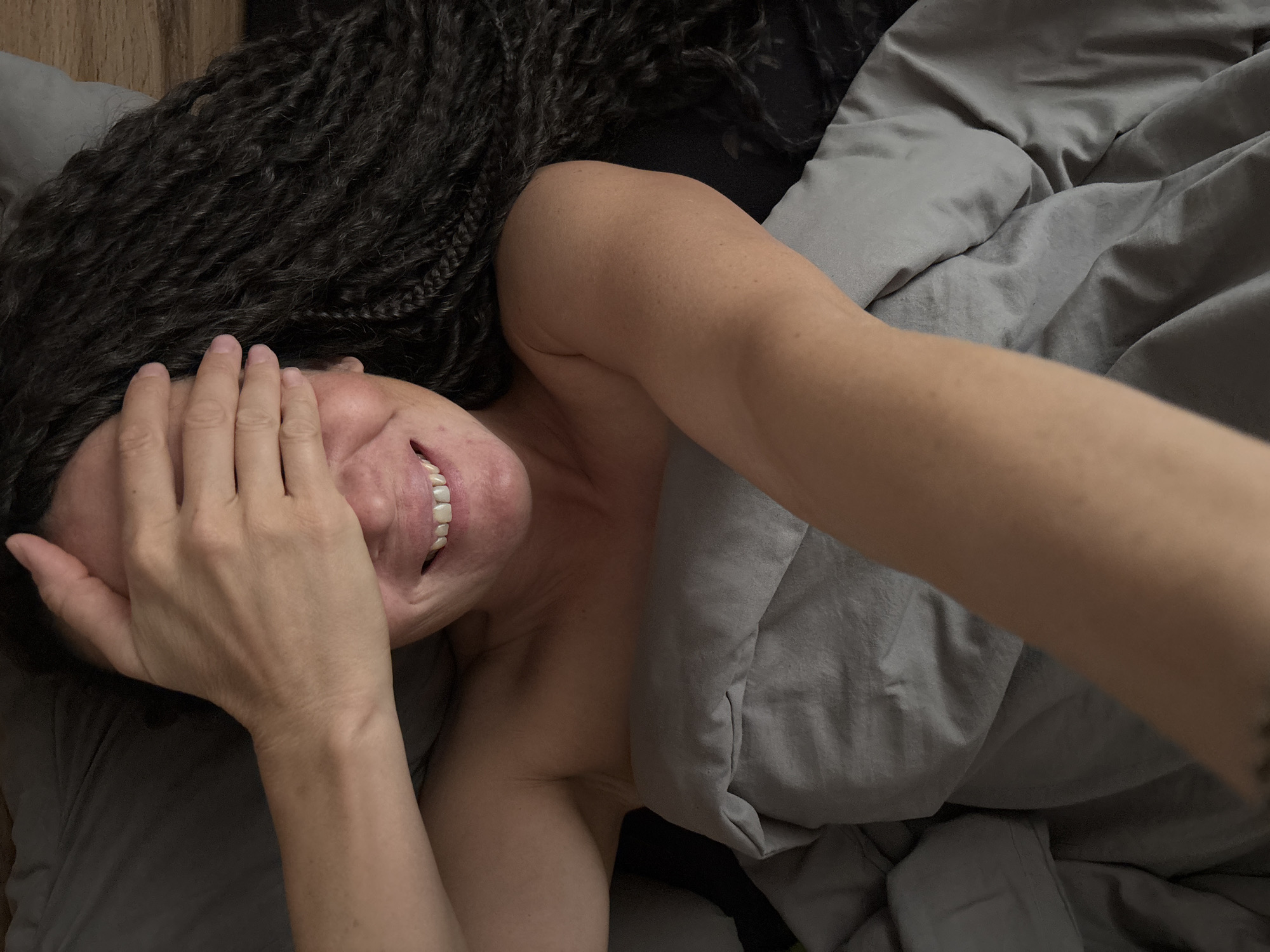 The image depicts an adult individual lying in a bed in the city of Chaikovsky, Russia. The person has a medium-dark skin tone and long, dark, textured hair, styled in braids or twists, which extends across the dark pillow behind their head. Their right hand is positioned over their upper face, covering the eyes and forehead. A wide smile is visible on the lower face, revealing white, even teeth. The individual's shoulders and upper chest are exposed, rising from beneath a light gray duvet or blanket that covers the rest of their body. A bare forearm extends into the lower portion of the frame, suggesting the image is a selfie. The scene is set indoors, likely in a bedroom, with a subdued lighting condition.