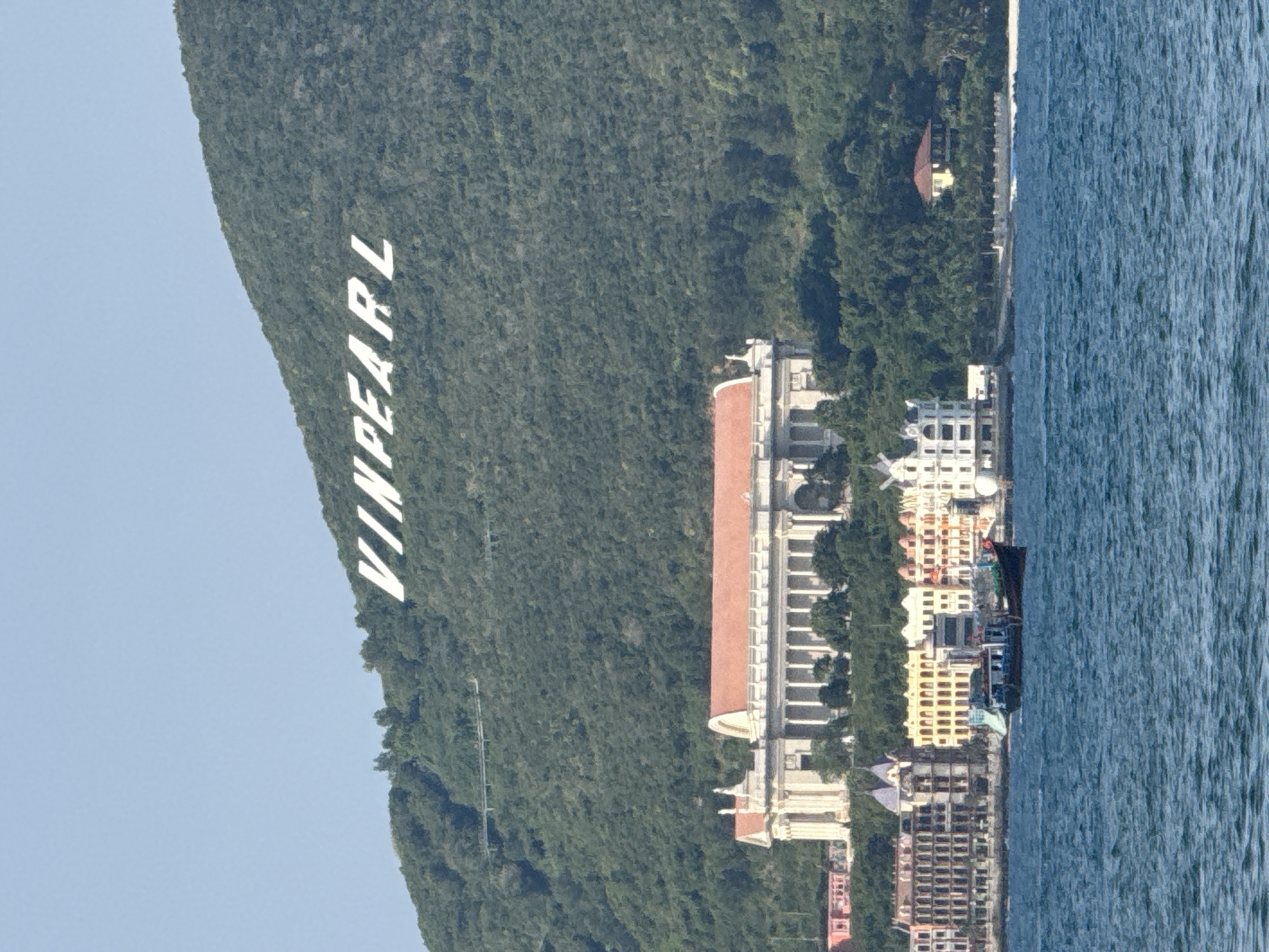 The image shows a daytime view of the coast of Nam Nha Trang Ward, Vietnam. In the background, large white letters spelling "VINPEARL" are visible on a green, tree-covered mountainside. Below the letters, a long, classical-style building with a red roof and white columns is situated. In the foreground, a body of blue water, likely the sea, is shown with a small dark boat on it. Along the shoreline, a row of colorful buildings, some with ornate architecture, are visible, hinting at a resort or entertainment complex. The overall scene suggests a tourist destination.