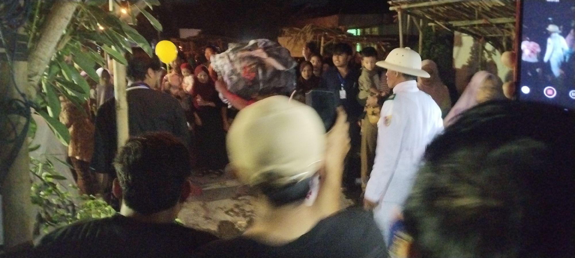 The image shows a nighttime outdoor gathering of people in Keting, Indonesia. The scene appears to be a street or open area with some foliage on the left and a structure with a thatched roof in the background. A group of people are visible, some are blurred due to motion. In the foreground, the back of two individuals wearing hats is prominent, suggesting they are looking at an event.
A central subject is a person carrying a large, fabric-covered object on their head, which is likely part of a cultural performance or ritual. This person is wearing a white uniform with a hat. Other individuals in the background are dressed in casual attire, and some are wearing headscarves. A bright yellow balloon is visible in the upper left quadrant of the image, adding a festive element. A smartphone with its screen on is held up in the right side of the frame, capturing the event. The overall atmosphere suggests a community event or celebration taking place at night.