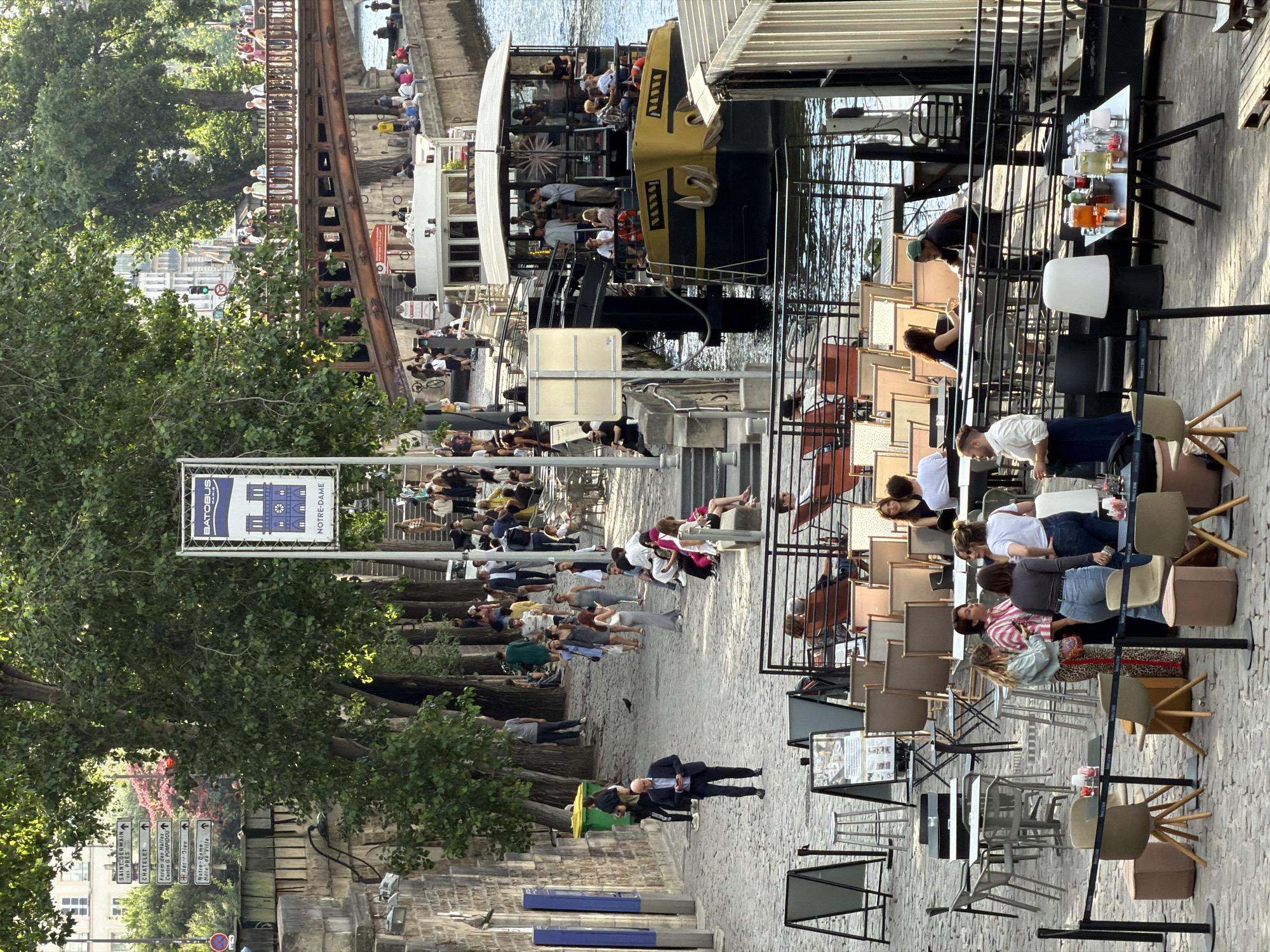 This is a vertical, eye-level, full shot of a busy scene along a riverbank in Paris, France, likely in the late afternoon due to the warm sunlight casting long shadows. On the left, a tree-lined promenade with a cobblestone path is filled with people walking and seated at tables. The trees are lush and green, suggesting a warm season.
In the middle ground, a yellow and black tourist boat, the BATOBUS, is docked. Passengers are visible on its deck. The river is calm and reflects the surrounding structures. Signs are posted, one prominently displaying "BATOBUS" with an image of Notre-Dame Cathedral and text indicating "NOTRE-DAME." Other directional signs point towards Chatelet, Hotel Dieu, and Notre-Dame, confirming the location.
To the right, an outdoor seating area with numerous tables and chairs is set up, with some people seated and others standing. Drinks and bottles are visible on one of the tables. The architecture in the background suggests a European city. The overall atmosphere is one of a lively, popular tourist spot with people enjoying the waterfront.