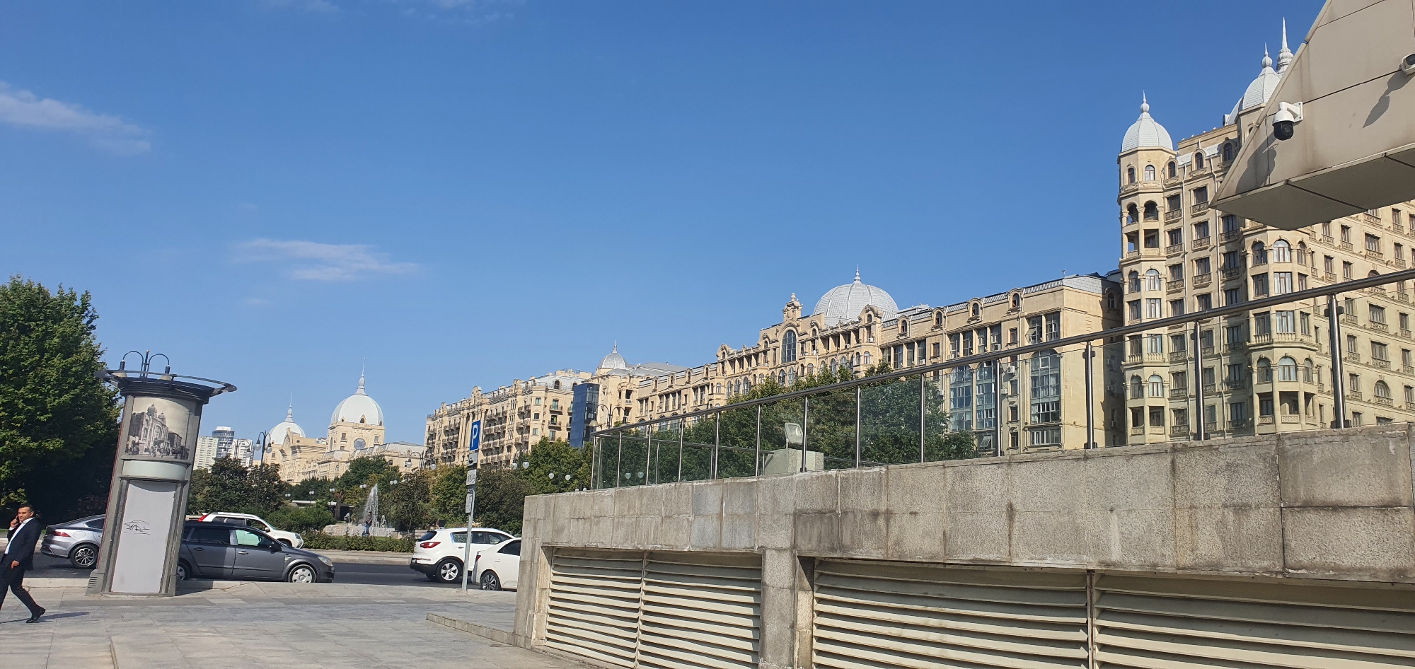 This image captures a street scene in Baku, Azerbaijan, focusing on a row of elegant, historic buildings with distinctive architectural features such as domes and ornate facades. The buildings are light yellow in color and appear to be multiple stories high. In the foreground, a concrete barrier with a slatted lower section runs horizontally across the frame. Behind this barrier, a glass railing outlines a raised walkway or structure. To the left, a tall, cylindrical monument with a curved top displays a historical image. Several cars are parked along the street and on the road. A man in a suit is walking on the sidewalk to the left, engaged in a phone conversation. The sky is a clear, bright blue with a few wispy clouds, suggesting a sunny day. Lush green trees are visible behind the buildings and along the street, contributing to the urban landscape. The overall impression is one of a well-maintained, historic city district on a pleasant day.
